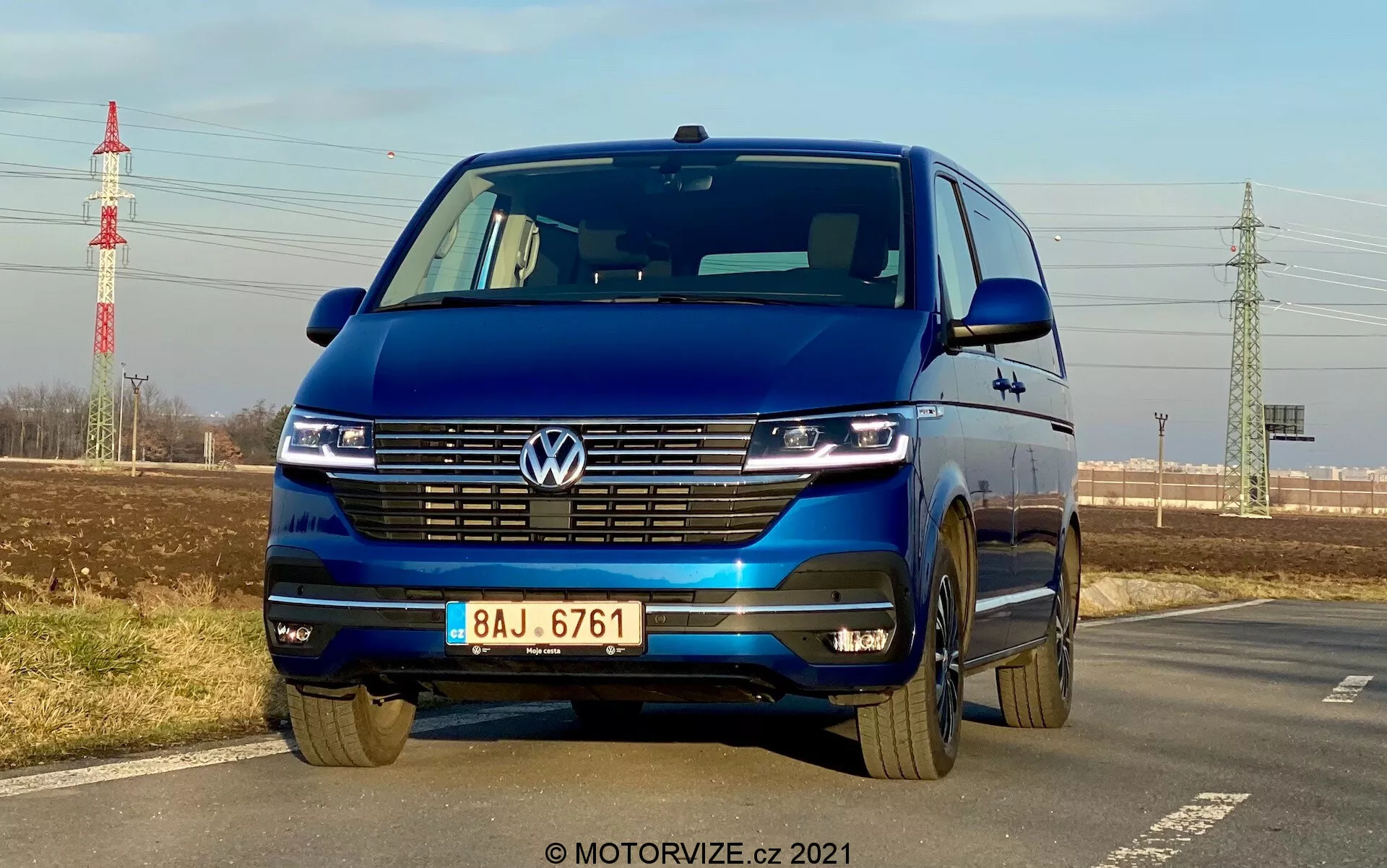 Niebieski Volkswagen T6-1 Multivan 2019 uchwycony z niskiego przedniego kąta podkreślającego grill, reflektory LED z technologią świateł dziennych, emblemat Volkswagena, szeroki grill, dwukolorowy zderzak i aluminiowe felgi; zaparkowany przy drodze z czystym niebem, odległą zielenią, słupem energetycznym i infrastrukturą w tle.