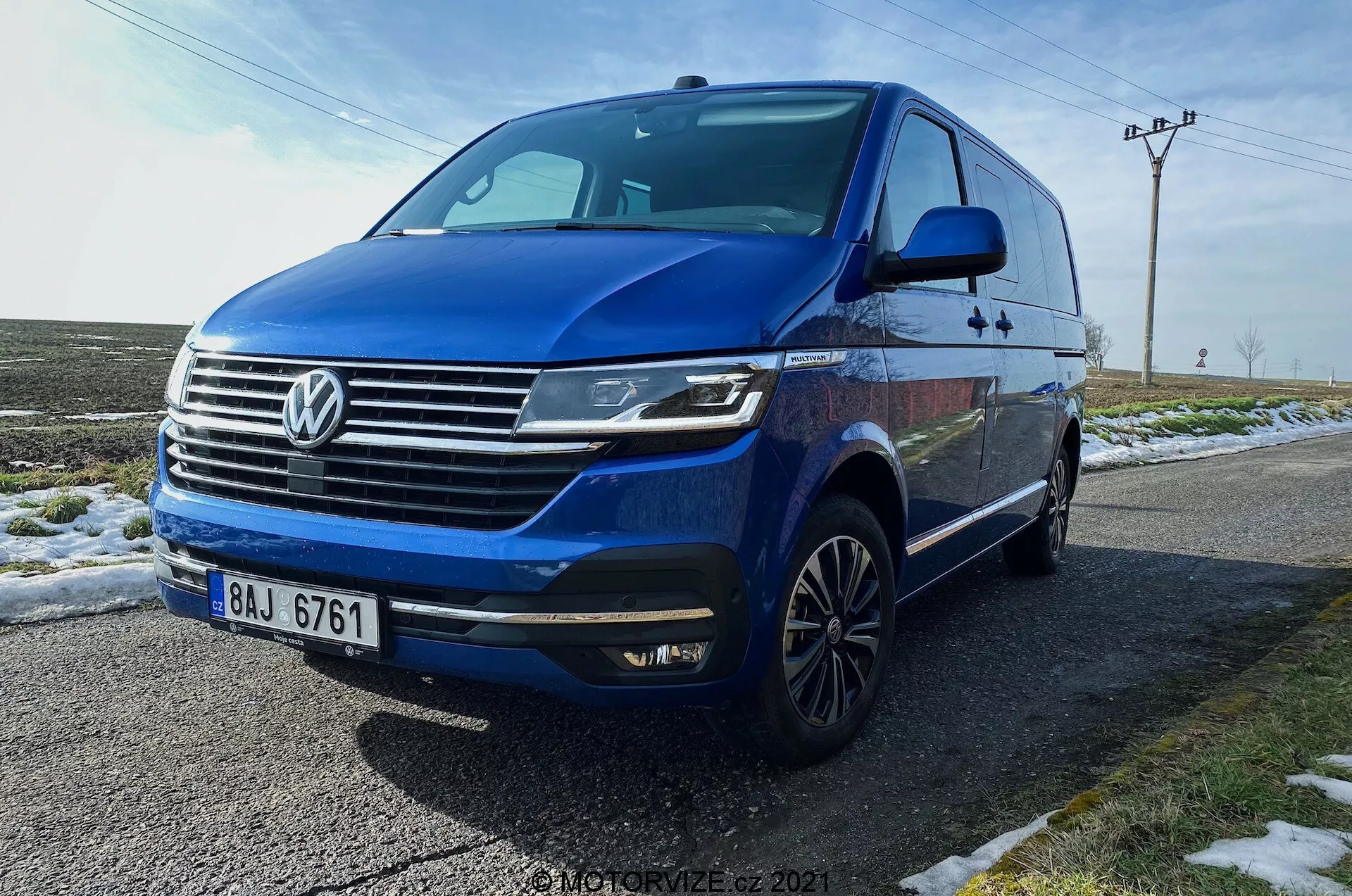 Niebieski Volkswagen T6.1 Multivan Transporter 2019 uchwycony z niskiego kąta trzech czwartych przodu, ukazujący przedni pas samochodu, grill z wyraźnym logo Volkswagena, poziome chromowane listwy, kanciaste reflektory LED z światłami do jazdy dziennej, przedni zderzak z zintegrowanymi światłami przeciwmgielnymi i wlotami powietrza, dynamiczna linia biegnąca wzdłuż boków, wieloramienne felgi aluminiowe, zintegrowane kierunkowskazy w lusterkach bocznych, przyciemniane szyby. Pojazd zaparkowany na mokrej drodze obok zaśnieżonej trawy, z wiejskim tłem z słupami elektrycznymi i otwartym niebem.
