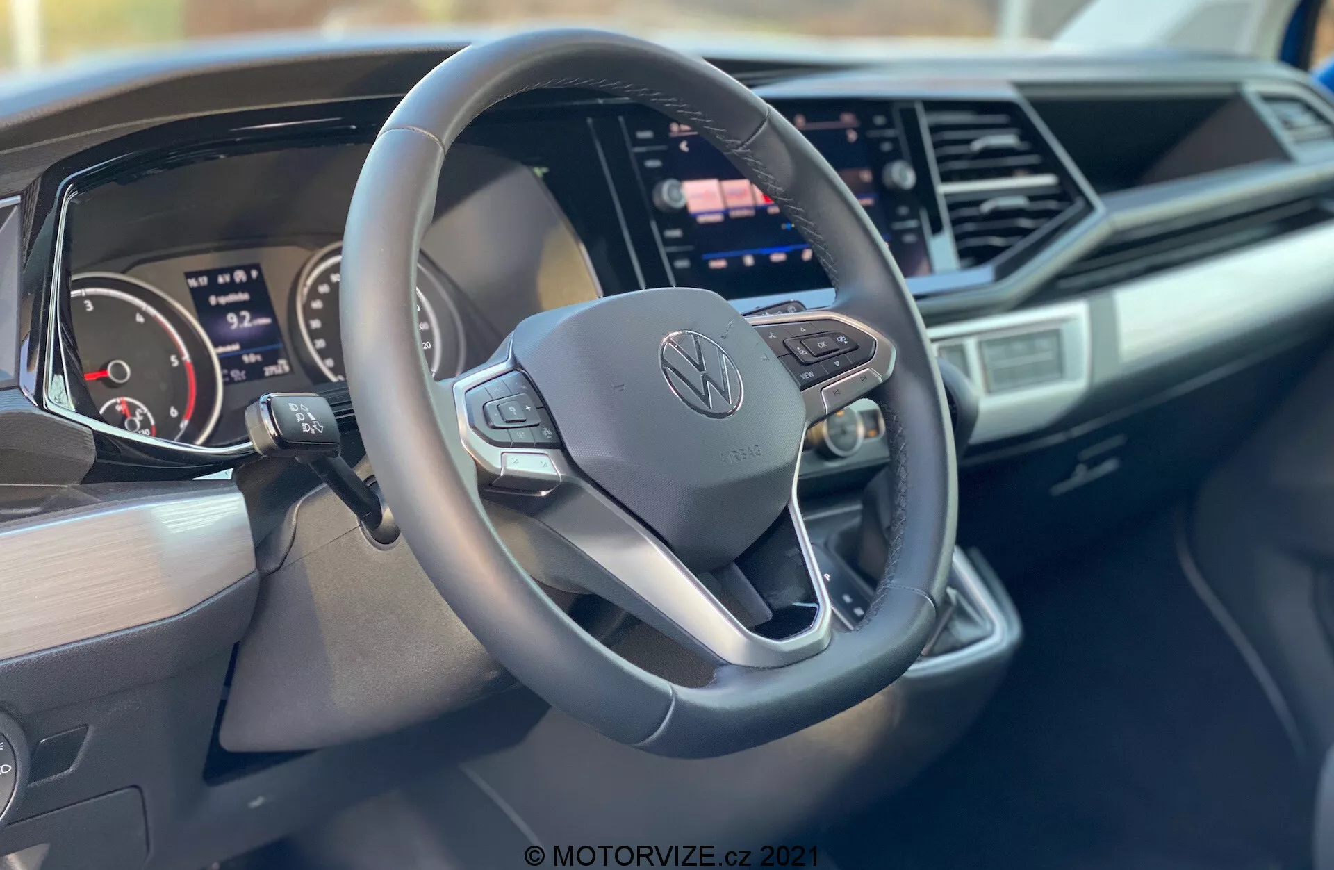 Zdjęcie wnętrza Volkswagen Nutzfahrzeuge Transporter 2019 (T6-1 Multivan) skupiające się na kierownicy i desce rozdzielczej. Zbliżenie wykonane z perspektywy pasażera przedstawia trójramienną, spłaszczoną u dołu kierownicę z przyciskami sterowania i centralnym logo Volkswagena, cyfrowy zestaw wskaźników, duży centralny ekran dotykowy systemu multimedialnego, elegancki projekt deski rozdzielczej z kanciastymi akcentami oraz oznakowanie marki po stronie pasażera.