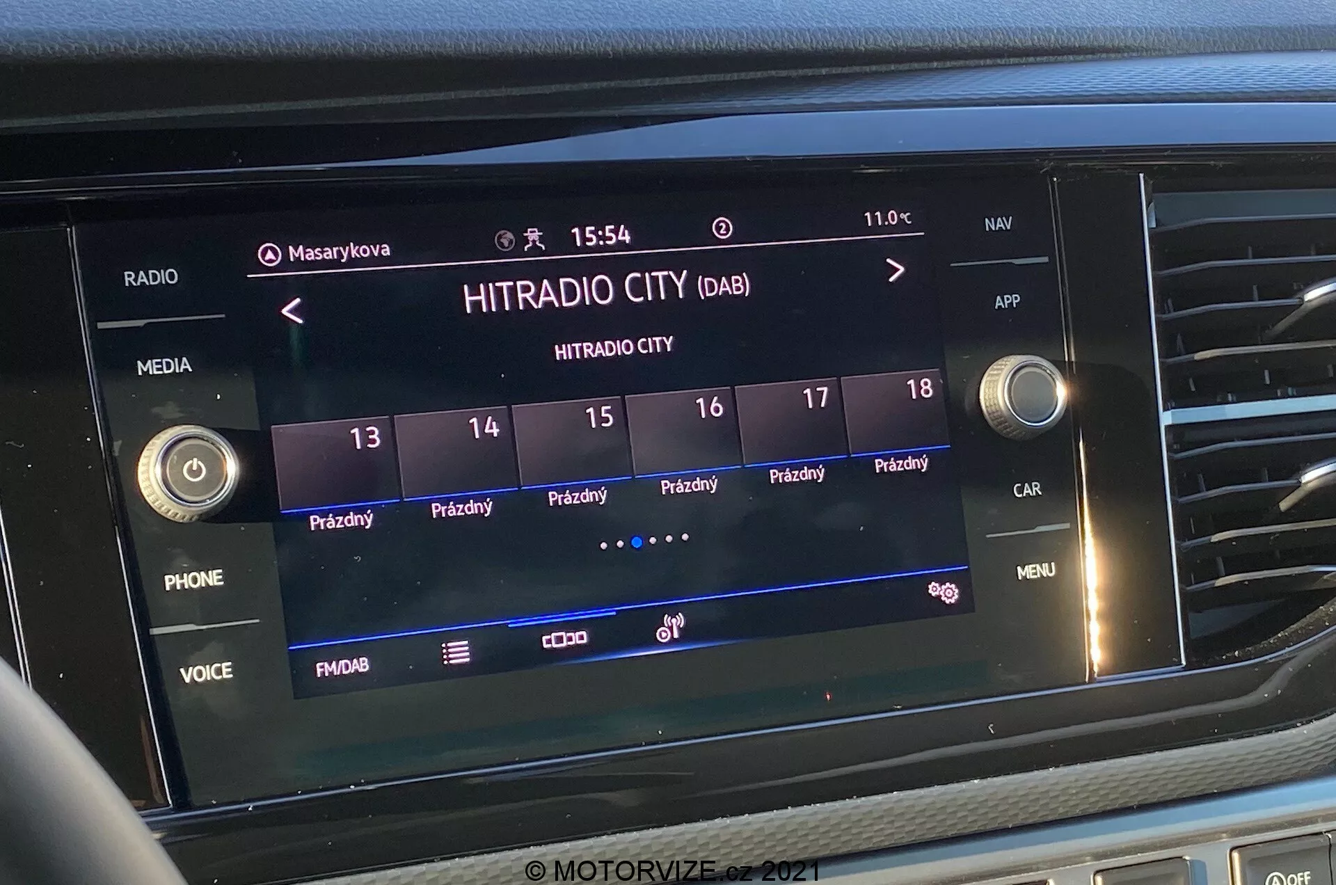 Zbliżenie przedniego widoku ekranu systemu infotainment w VW-Nutzfahrzeuge Transporter 2019 (T6-1 Multivan). Obraz przedstawia wybraną stację radiową 'HITRADIO CITY' z różnymi opcjami dotykowymi i przyciskami funkcji jak 'PHONE', 'VOICE' i 'MENU'. Wyświetlacz pokazuje godzinę '15:54' i zewnętrzną temperaturę '11.0°C' z ikonami profili użytkowników, ustawieniami pojazdu oraz siłą sygnału. Ekran jest zintegrowany w centralnej części deski rozdzielczej, otoczony przyciskami dotykowymi i pokrętłami sterującymi.