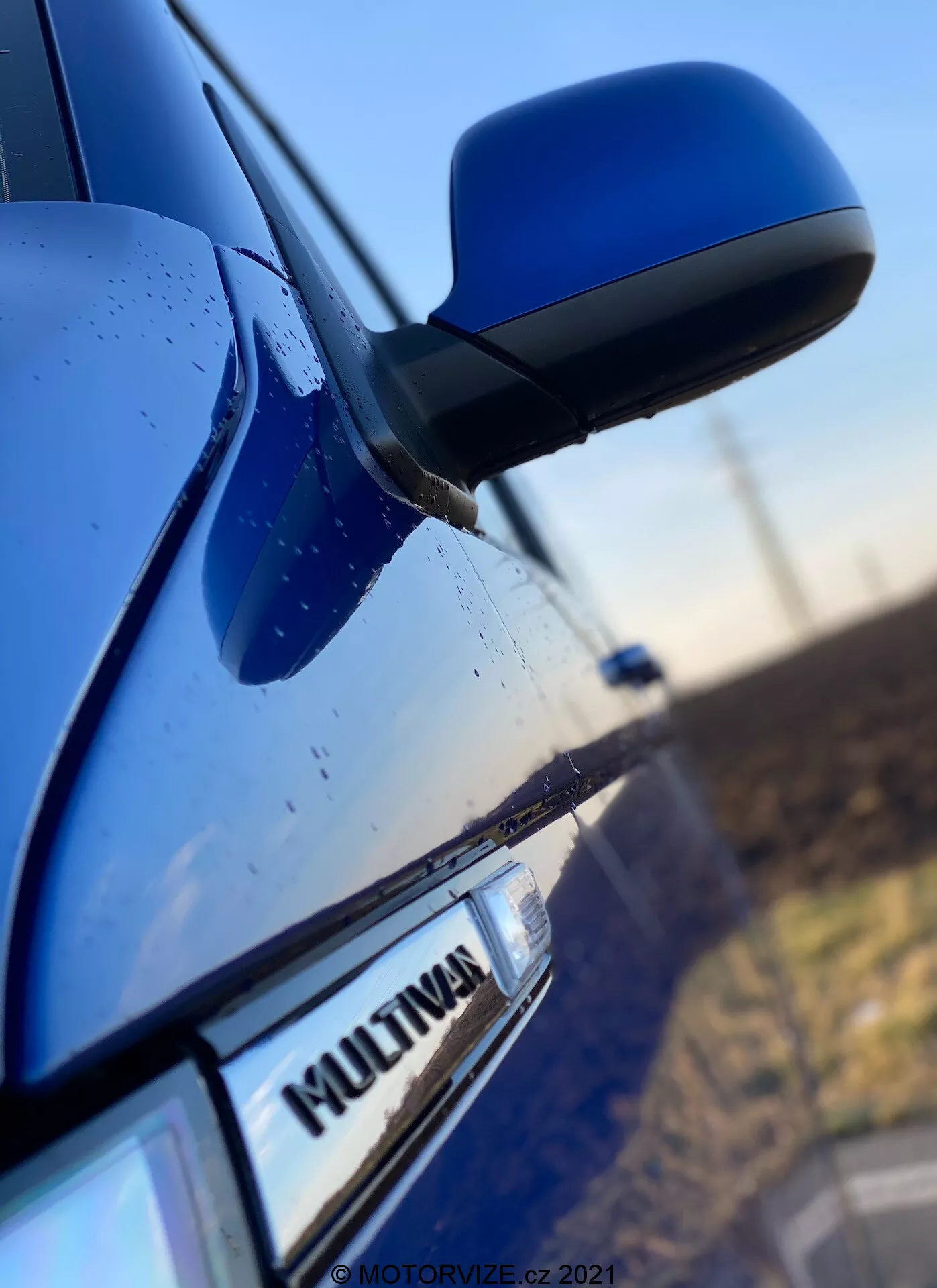 Zbliżenie na bok niebieskiego VW Nutzfahrzeuge Transporter 2019 (T6-1-Multivan) ukazujące część drzwi pasażera przedniego, lusterko boczne i fragment drzwi tylnych. Uwaga skupiona jest na odznace 'MULTIVAN' na samochodzie, z lekko rozmytym, aerodynamicznym lusterkiem bocznym. Obudowa lusterka pasuje do koloru nadwozia, a widoczne krople wody na powierzchni sugerują właściwości wodoodporne lub niedawną ulewę.