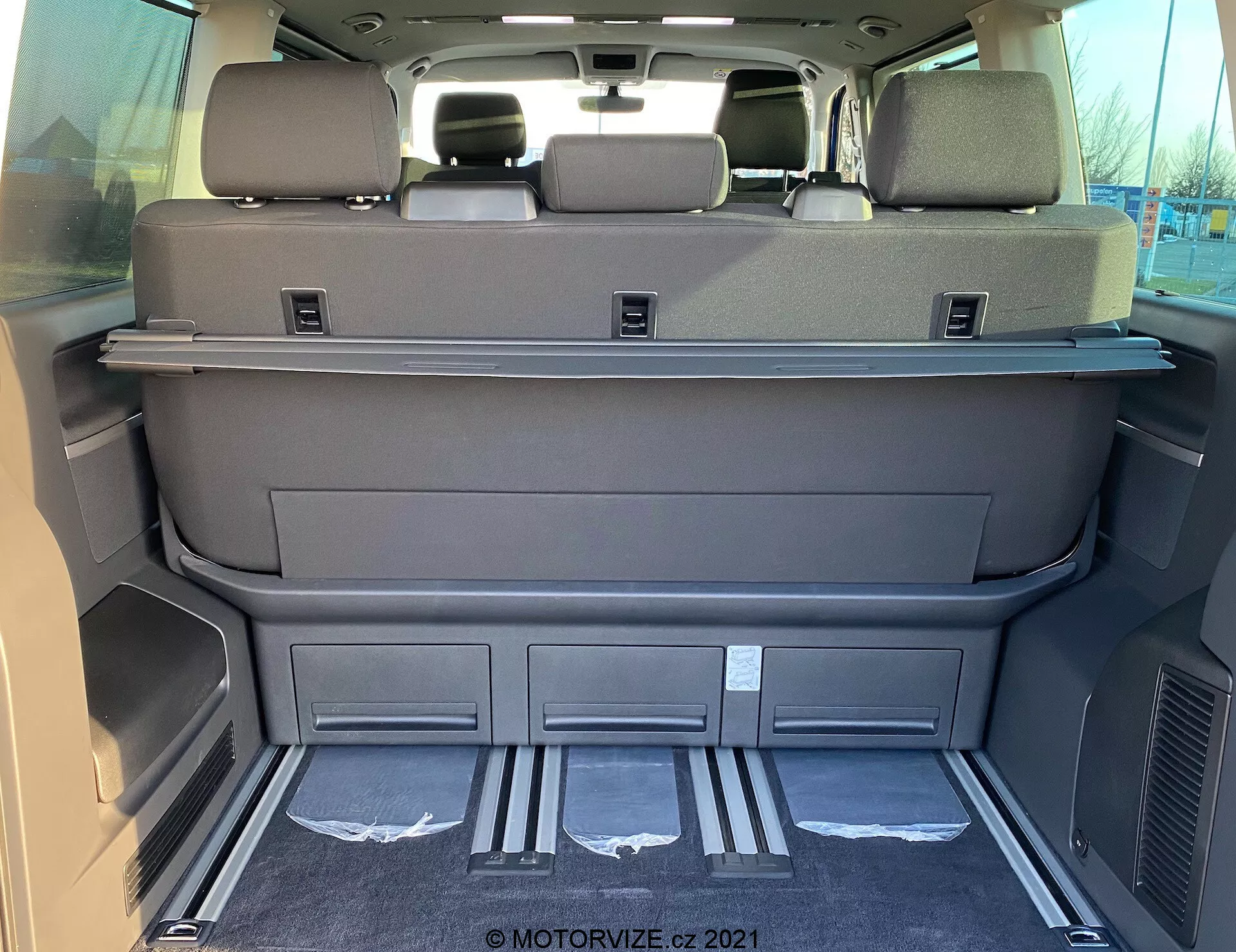 Wnętrze przestrzeni ładunkowej samochodu VW Nutzfahrzeuge Transporter T6.1 Multivan 2019 widziane od tyłu, ukazujące przestronną przestrzeń z złożonymi siedzeniami trzeciego rzędu, w kolorze ciemnoszarym lub czarnym, półkę osłaniającą ładunek, zagłówki na tylnych siedzeniach, metalowe osłony na krawędzi przestrzeni, ukształtowane schowki, kratki wentylacyjne, czysty dywanik i możliwe elektroniczne sterowanie siedzeniami lub tylną klapą.