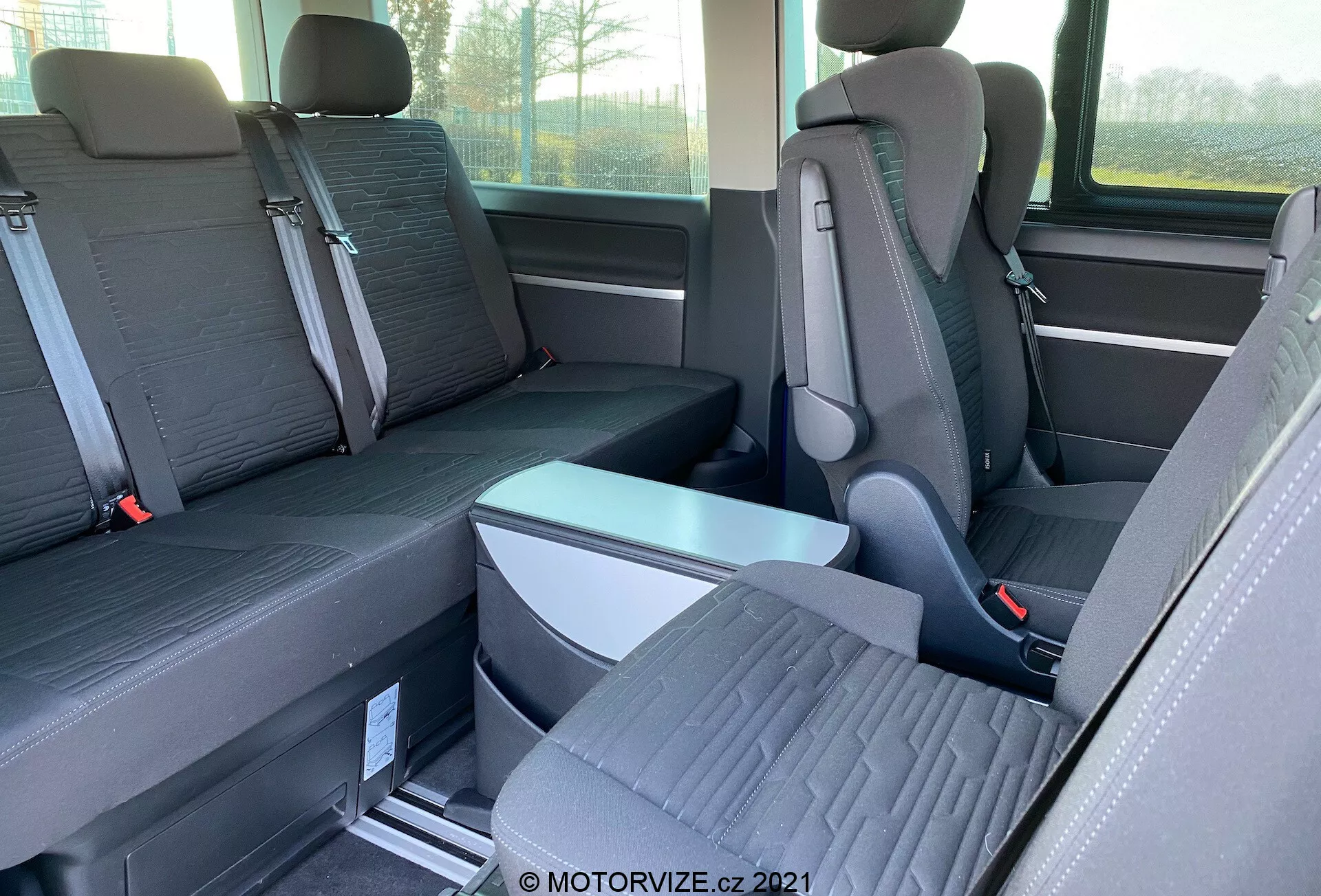 Widok wnętrza VW-Nutzfahrzeuge Transporter 2019 (T6.1 Multivan) skoncentrowany na siedzeniach drugiego i trzeciego rzędu. Fotografowane z boku na poziomie oczu, zdjęcie ukazuje indywidualne siedzenia z widocznymi zagłówkami w drugim rzędzie oraz ławkę dla trzech pasażerów w trzecim rzędzie. Siedzenia są dwukolorowe, a podłoga zawiera metalowe szyny do przestawiania siedzeń. Widać składany podłokietnik i klamry pasów bezpieczeństwa. Widać także częściowo okno i boczny schowek.