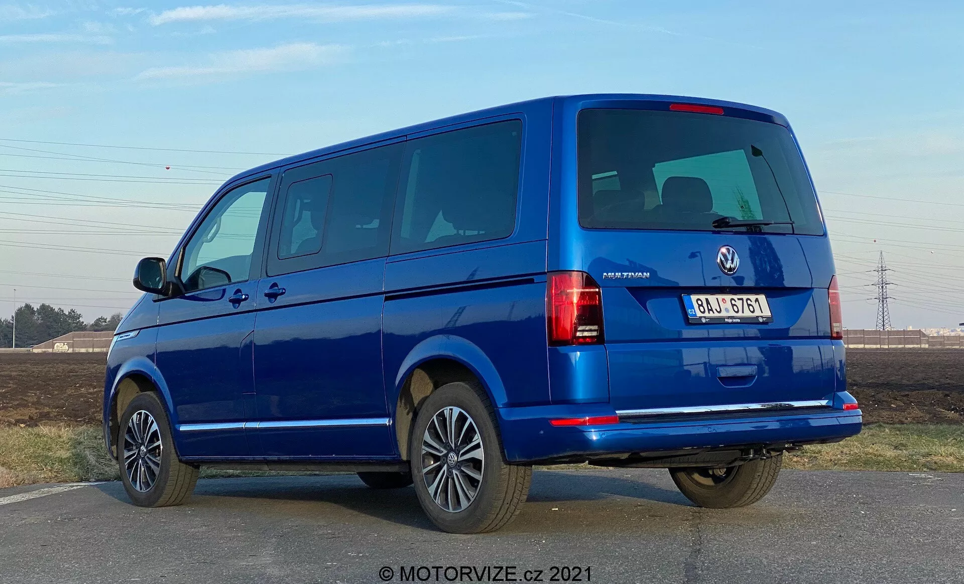 Niebieski VW-Nutzfahrzeuge Transporter 2019 (T6-1 Multivan) widziany z perspektywy trzech czwartych od tyłu. Obraz ukazuje tylne klosze, tylne okna i klapę tylną oraz profil boczny z drzwiami i oknami pasażerskimi. Furgonetka zajmuje dużą część kadru na poziomie oczu, ukazując dwuskrzydłowe tylne drzwi i kilka bocznych okien, co wskazuje na model pasażerski. Aluminiowe felgi, zderzaki w kolorze nadwozia i lusterka boczne sugerują wysoki poziom wyposażenia. Oświetlenie sugeruje, że zdjęcie wykonano w dzień przy bezchmurnym niebie, a pod pojazdem znajdują się wyraźne cienie.