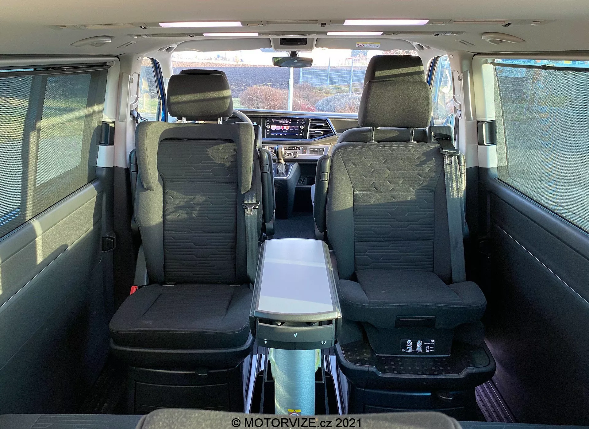 Wnętrze VW Nutzfahrzeuge Transporter T6-1 Multivan 2019 widziane od tyłu, ukazujące skórzane fotele, podłokietniki w drugim rzędzie, składany stolik, panoramiczny dach, indywidualne wyloty powietrza i zaawansowany system infotainment; podkreśla komfort pasażerów i przestronną kabinę.