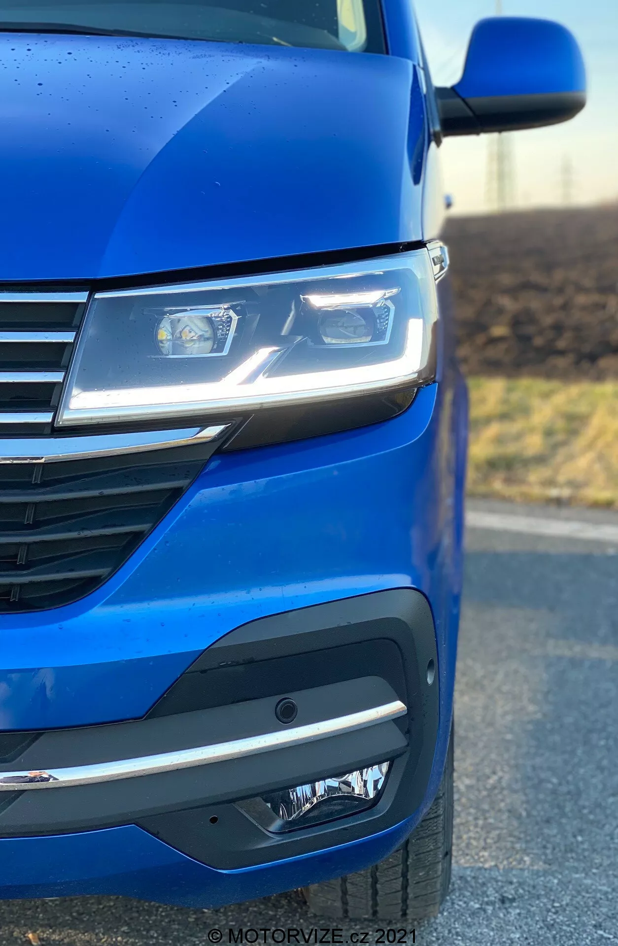 Zbliżenie na przednią lewą stronę VW Nutzfahrzeuge Transporter 2019 (T6.1 Multivan), podkreślające metaliczny niebieski lakier, zaawansowane reflektory LED, nowoczesny design grilla z poziomymi liniami oraz wyrafinowany przedni zderzak z halogenami lub jednostkami czujników. Kąt zdjęcia podkreśla aerodynamikę i szczegóły stylistyczne samochodu.
