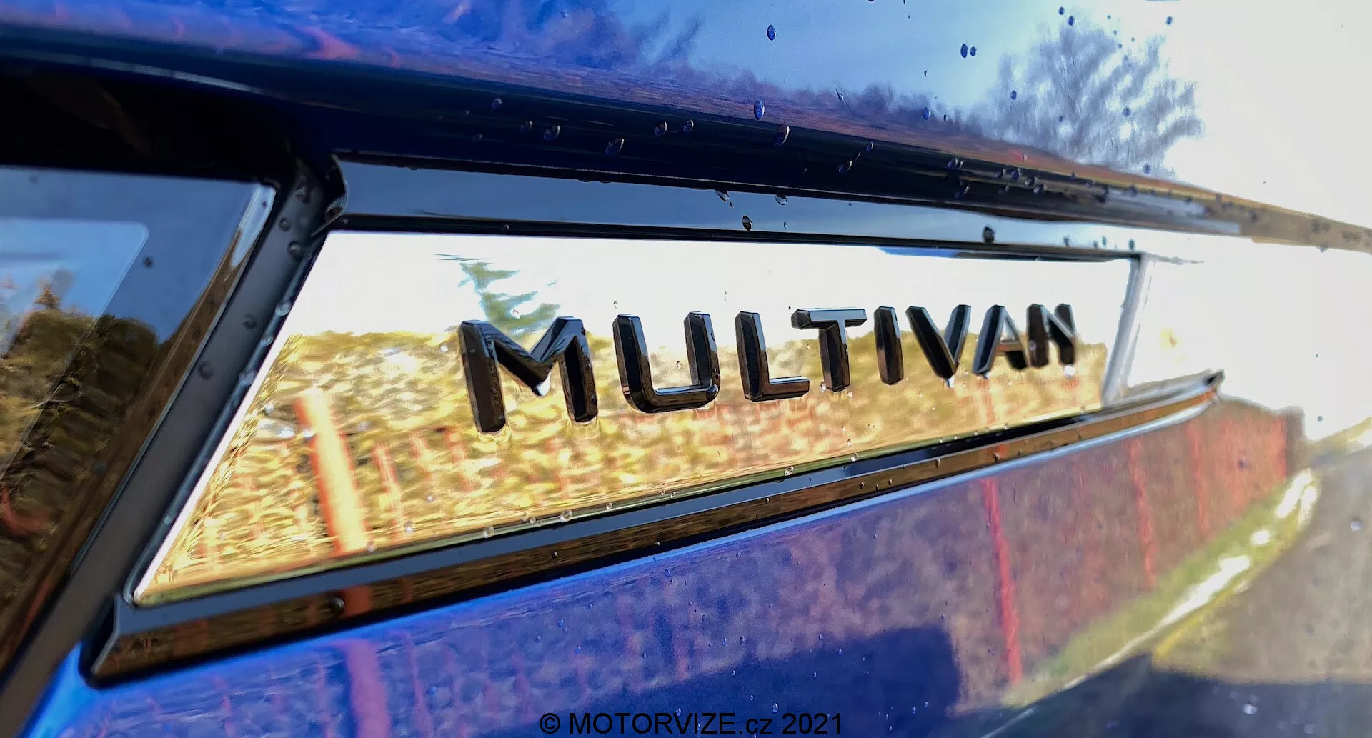 Zbliżenie części VW Nutzfahrzeuge Transporter 2019 (T6-1 Multivan) z naciskiem na znaczek 'MULTIVAN'. Zdjęcie jest wykonane z niskiego tylniego boku, patrząc lekko w górę, gdzie tylna ćwiartka okna pojazdu spotyka się z krawędzią dachu. Lekko widoczne odbicia na szybie okna sugerują plener. To ciasno wykadrowane zdjęcie podkreśla emblematykę modelu producenta.
