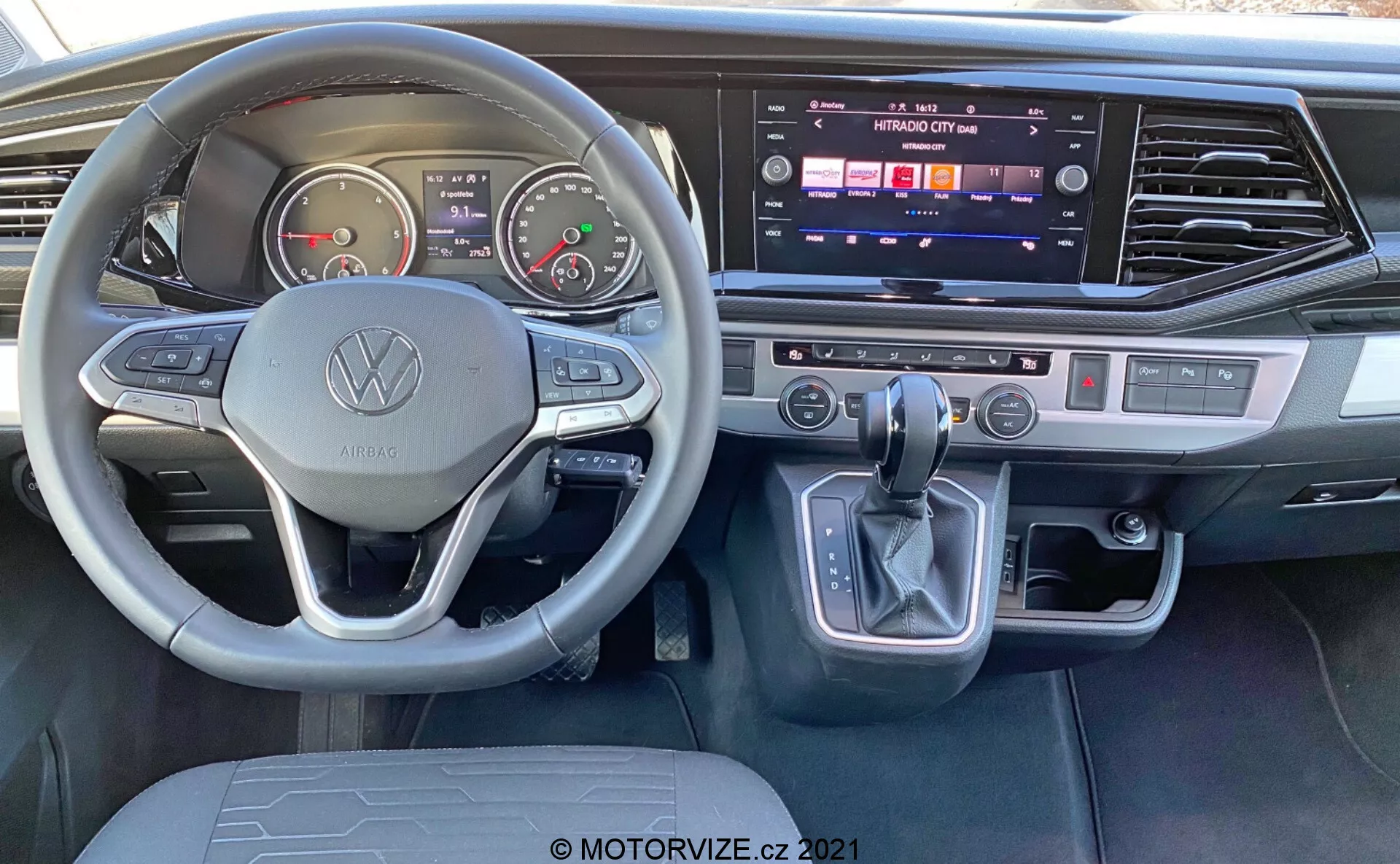 Widok wnętrza VW Nutzfahrzeuge Transporter 2019 T6.1 Multivan z perspektywy miejsca kierowcy. Zawiera kierownicę z logo Volkswagen i przyciskami wielofunkcyjnymi, analogowe zegary, wyświetlacz cyfrowy, konsolę środkową z aktywnym dotykowym systemem informacyjno-rozrywkowym, dwustrefową klimatyzację, dźwignię zmiany biegów z elektronicznym przyciskiem parkowania oraz nowoczesne akcenty z szczotkowanego metalu.