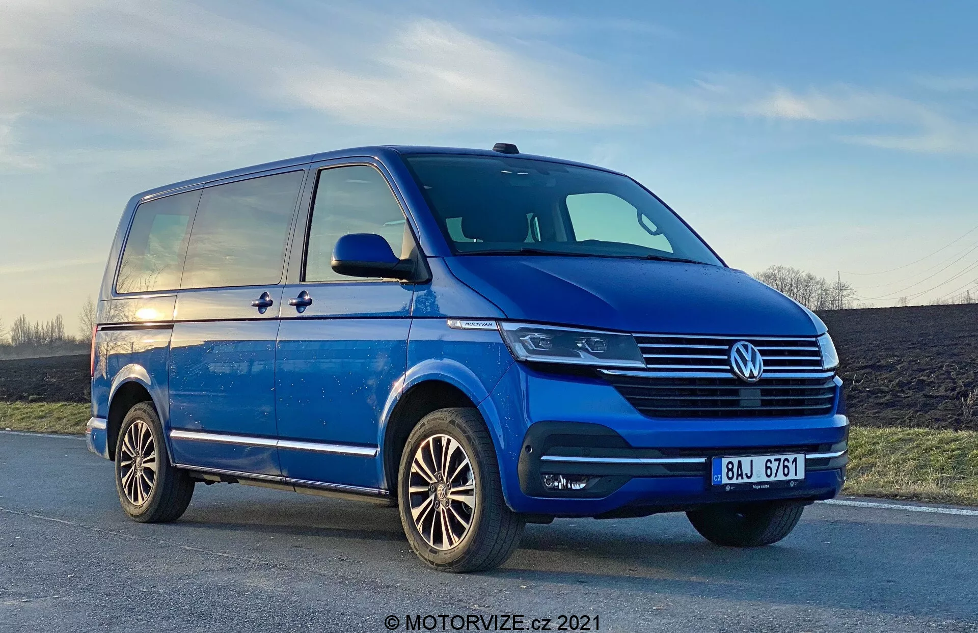 Niski widok błękitnego Volkswagena T6-1 Multivan 2019, ukazujący lewy przedni kwadrant z perspektywy trzy czwarte. Samochód jest zaparkowany na lekko nachylonej drodze, z wyraźnymi chromowanymi detalami na kracie, centralnym emblem VW i nowoczesnymi jednostkami reflektorów, które mogą wykorzystywać technologię LED. Światła przeciwmgielne są zintegrowane z zderzakiem, który ma także otwory wentylacyjne. Profil boczny ukazuje przesuwane drzwi, charakterystyczną linię rozciągającą się od przedniego błotnika do tylnego oraz masywne lusterka boczne z kierunkowskazami. Widzimy również felgi aluminiowe o wieloramiennym wzorze i dobrze utrzymany bieżnik opon. Niebo w tle pokazuje odcień zachodu słońca.