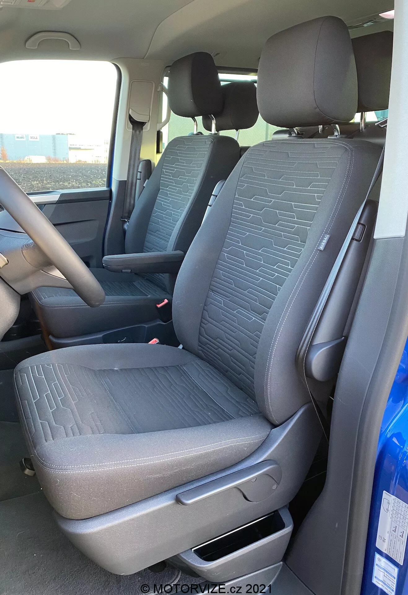 Wnętrze VW Nutzfahrzeuge Transporter 2019 T6.1 Multivan, ukazujące przednie siedzenie pasażera i część siedzenia kierowcy z perspektywy zewnętrznej po stronie pasażera. Zdjęcie zrobione pod lekko niskim kątem podkreśla teksturowaną tkaninę z geometrycznymi wzorami na siedzeniach, dźwignię regulacji manualnej, pasy bezpieczeństwa oraz część konsoli środkowej i kierownicy. Naturalne światło oświetla wnętrze pojazdu.