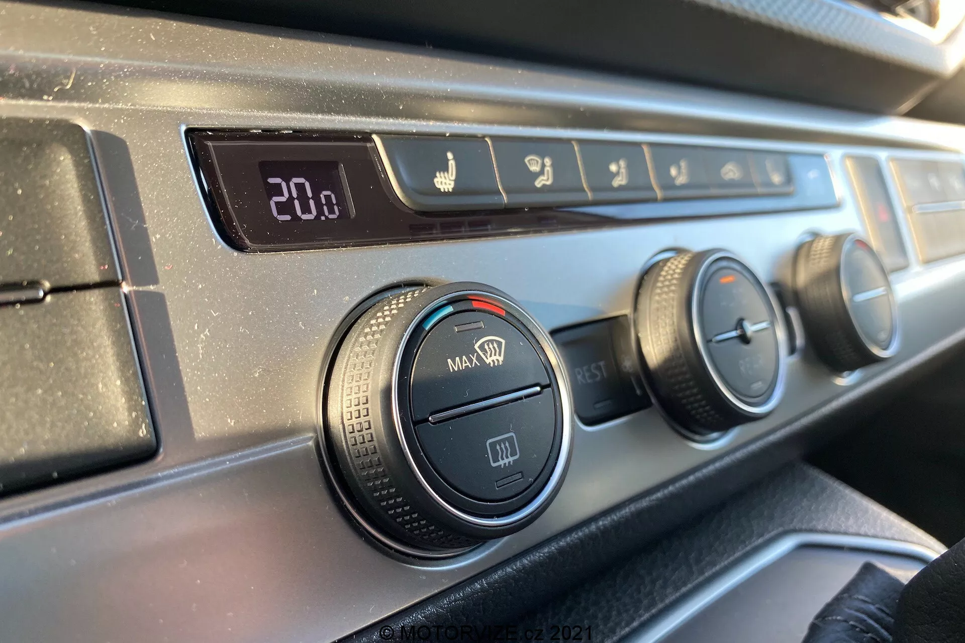 Zbliżenie panelu sterowania klimatyzacją na desce rozdzielczej VW Transporter T6.1 Multivan 2019 z perspektywy osoby siedzącej z przodu, z cyfrowym wyświetlaczem temperatury ustawionym na 20,0 stopni Celsjusza, sterowaniem kierunkiem przepływu powietrza, trzema pokrętłami do ustawiania klimatyzacji i wentylatora oraz przyciskiem recyrkulacji powietrza, wszystko z teksturowanym czarnym plastikiem i gładkim, ciemnym wykończeniem deski rozdzielczej.