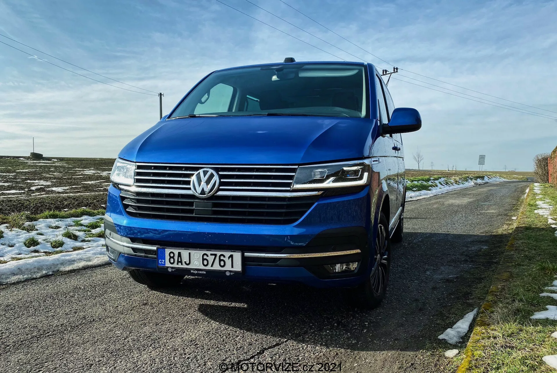 Niski widok z przedniego trzech czwartych perspektywy Volkswagena T6.1 Multivan 2019, z przednią kratką z poziomymi liniami, charakterystycznym logo VW w centrum kratki, reflektorami prawdopodobnie z LED DRL i bi-ksenonowymi lub LED światłami, i przednim zderzakiem z przeciwmgielnymi światłami i wlotami powietrza. Obraz pokazuje część lewego boku, w tym drzwi kierowcy i błotnik, z widoczną maską i przednią szybą. Samochód ma głęboki niebieski kolor i jest zaparkowany na wiejskiej drodze z odrobiną śniegu na ziemi i czystym niebieskim niebem powyżej.