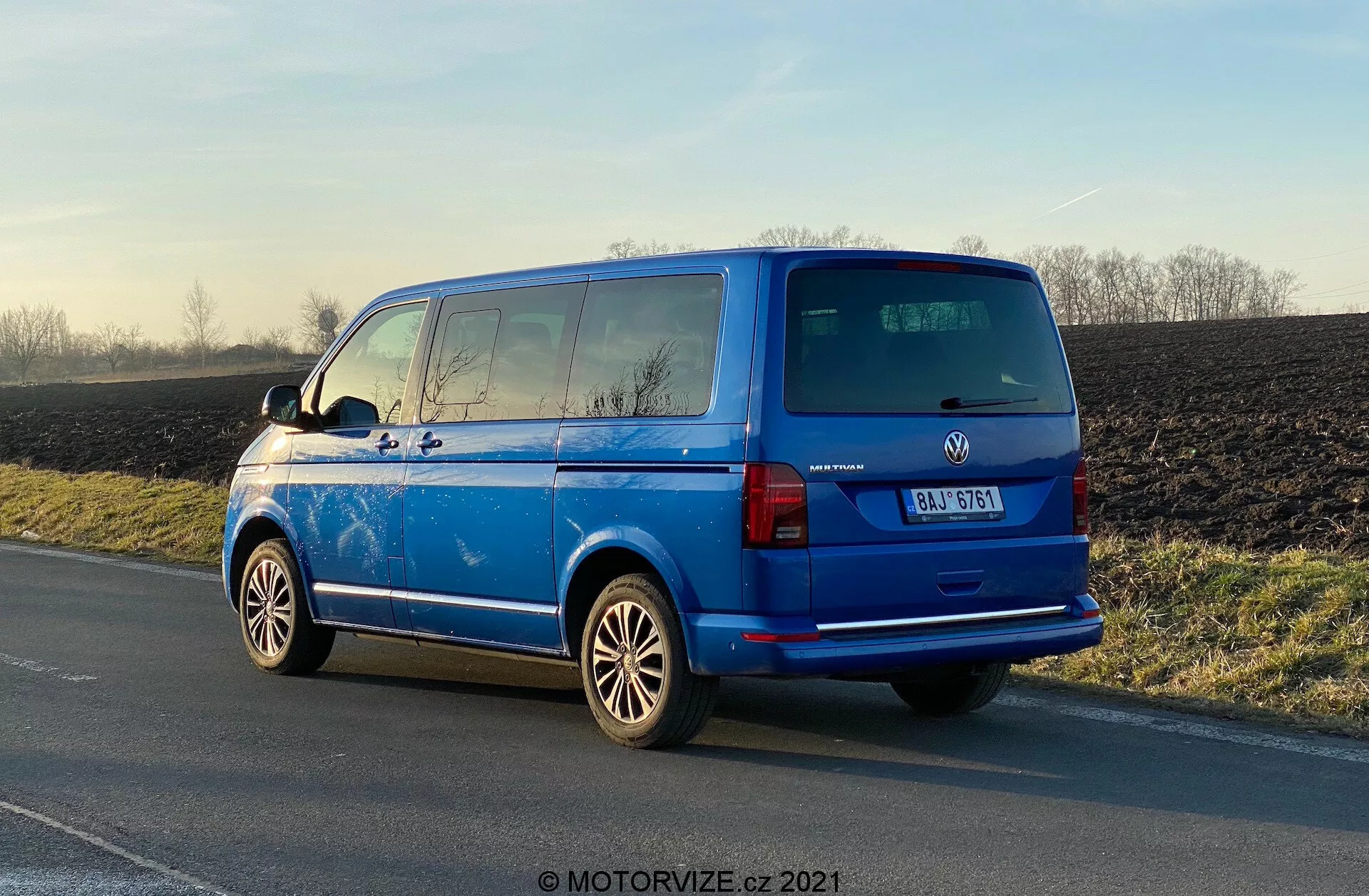 Intensywnie niebieski Volkswagen Transporter 2019 (T6-1 Multivan) zaparkowany na poboczu, widok z tyłu pod kątem trzech czwartych, pokazujący tylne światła, tylne okno, tylne drzwi, znaczek producenta, szczegóły modelu pojazdu; widoczne felgi aluminiowe, zewnętrzne lusterka boczne, linie odmrażacza tylnych szyb, prywatne przyciemniane szyby drugiego rzędu, antena na dachu; zaparkowany w naturalnym świetle dziennym z odbiciami podkreślającymi polerowaną powierzchnię, wiejskie tło.