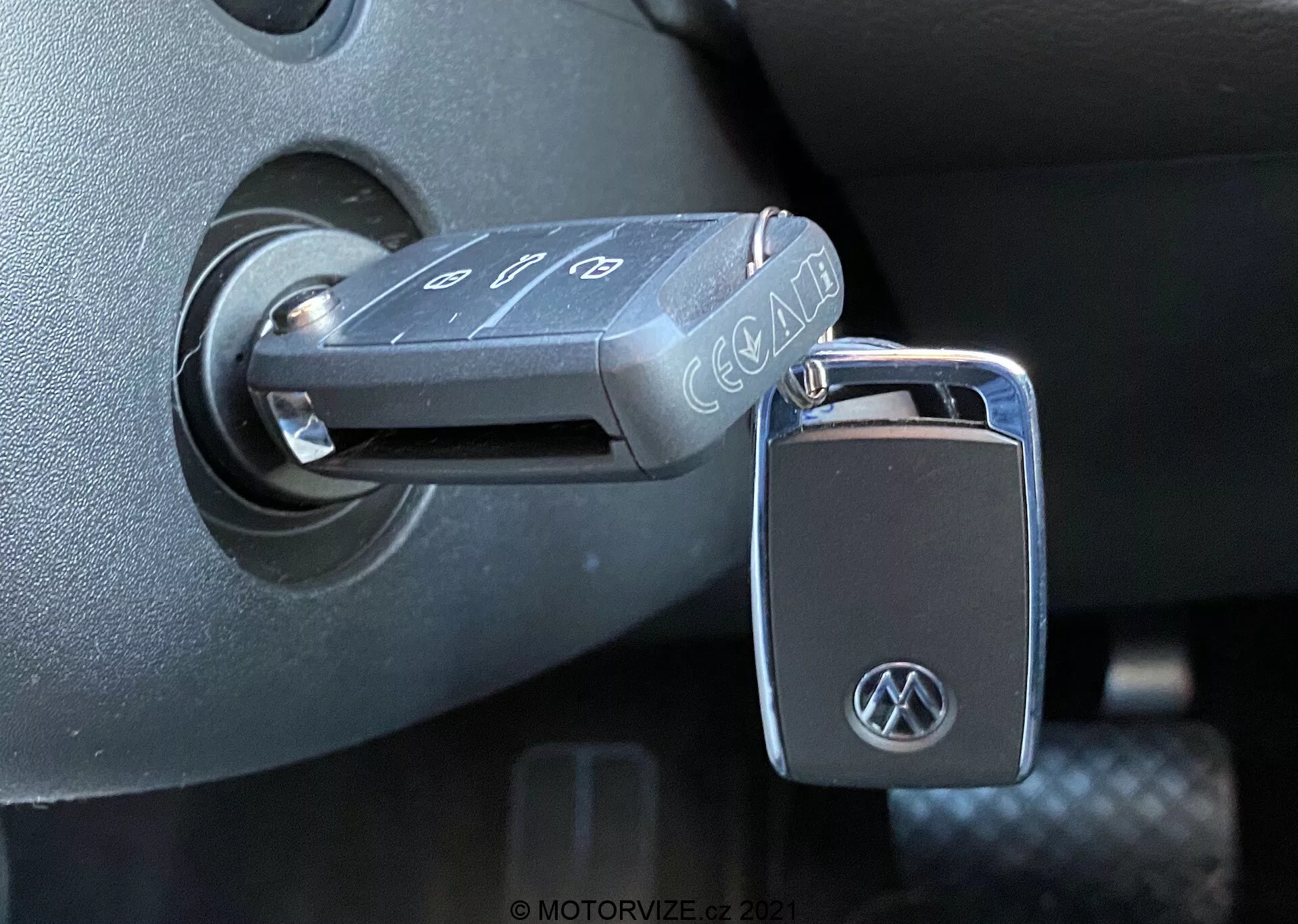 Zbliżenie na składany klucz Volkswagena włożony do stacyjki samochodu VW Nutzfahrzeuge T6-1 Multivan z 2019 roku. Klucz, z przyciskami do zamykania, otwierania i alarmu panicznego, ma logo VW na breloku. Stacyjka znajduje się w ciemnym plastikowym otoczeniu kolumny kierownicy, wskazując, że zapłon jest w pozycji 'off'. Zdjęcie, zrobione z bocznego kąta przy naturalnym świetle, podkreśla szczegółowe tekstury i wykończenia wnętrza pojazdu.
