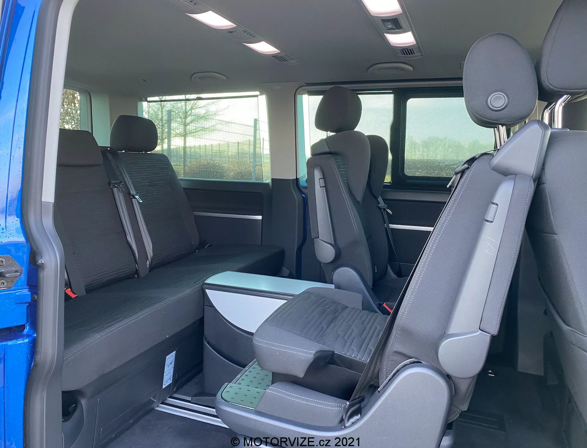 Wnętrze VW Transporter T6.1 Multivan 2019 ukazujące drugi i trzeci rząd siedzeń z perspektywy tylnego siedzenia pasażera, z konfiguracją siedzeń 2-3-2 i ciemną tapicerką. Częściowy widok na zewnątrz przez otwarte drzwi, zagłówki na wszystkich siedzeniach, podniesione siedzenia w trzecim rzędzie dla lepszej widoczności.
