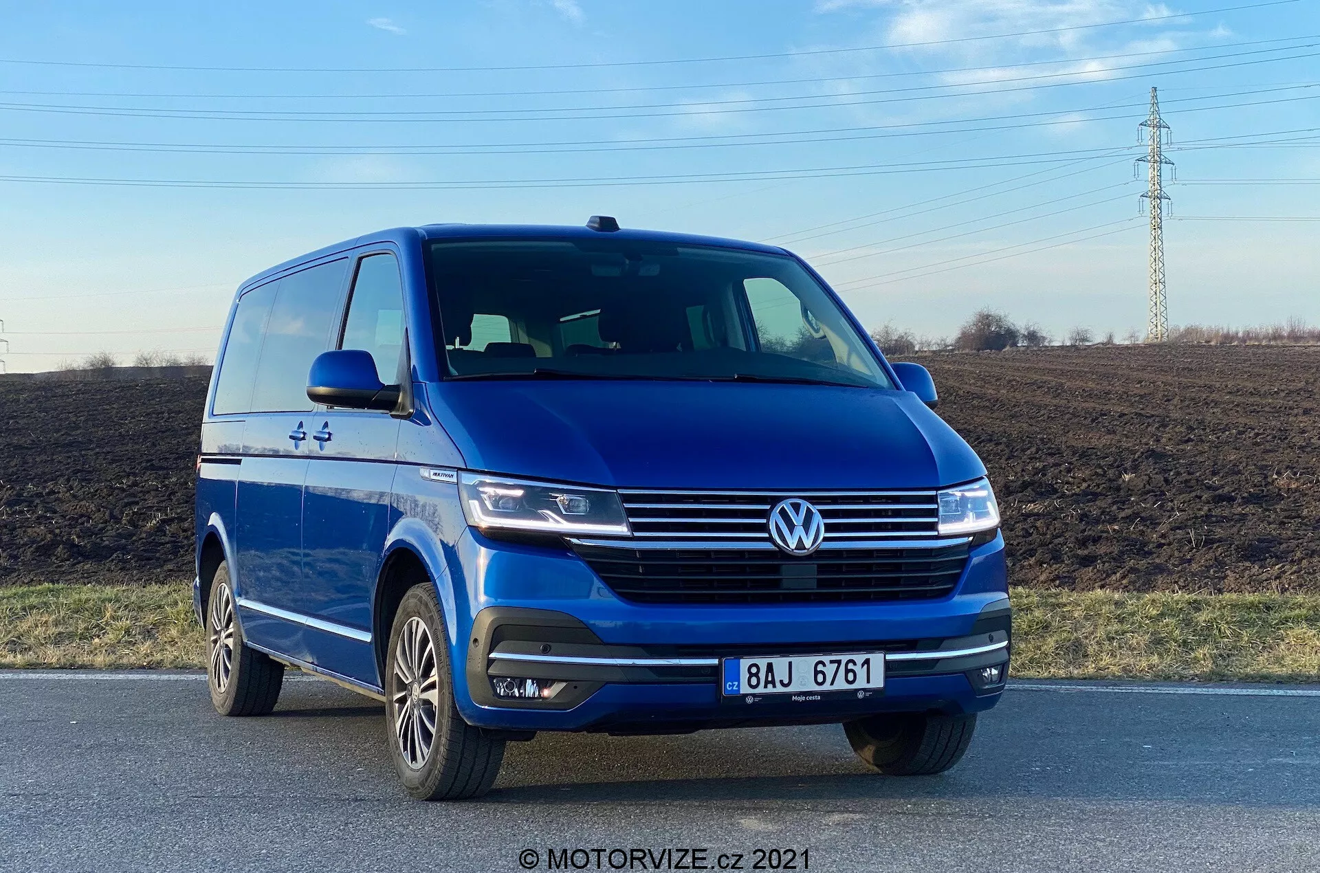 Zdjęcie przedstawia niebieski Volkswagen VW-Nutzfahrzeuge Transporter 2019 (T6-1 Multivan) uchwycony z niskiego kąta przedniego, ukazując przednią lewą ćwiartkę z wyraźnym widokiem na przednią fasadę i profilem strony kierowcy. Pojazd znajduje się na drodze, podkreślając takie elementy jak wyeksponowany znaczek Volkswagen, eleganckie reflektory, rzeźbione linie maski, zintegrowane światła przeciwmgielne w zderzaku oraz wieloramienne felgi aluminiowe. Zdjęcie wykonane w świetle dziennym podkreśla stylizację zewnętrzną vana i technologie wspomagające kierowcę, takie jak czujniki parkowania.