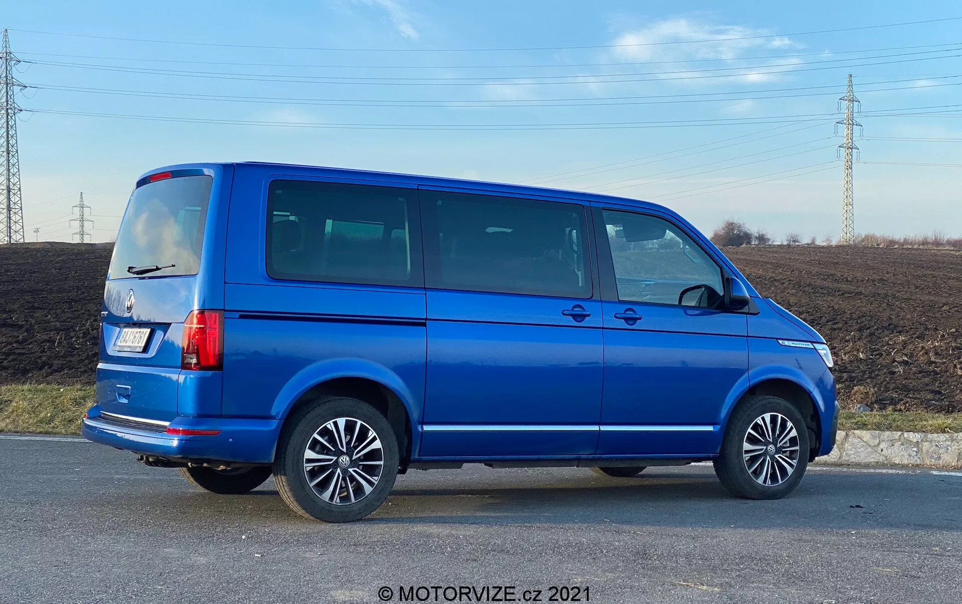 Pełny widok z profilu bocznego niebieskiego VW Nutzfahrzeuge Transporter z 2019 roku (T6.1 Multivan) z felgami aluminiowymi, widocznymi liniami drzwi, przyciemnianymi szybami bocznymi, kanciastą sylwetką, pionowymi światłami tylnymi, anteną na dachu i jasnoniebieskim metalicznym lakierem. Zdjęcie zrobiono pod kątem, co daje perspektywę na bok i tył pojazdu, ustawionego na obszarze wiejskim z polem i liniami wysokiego napięcia w tle, w świetle dziennym.