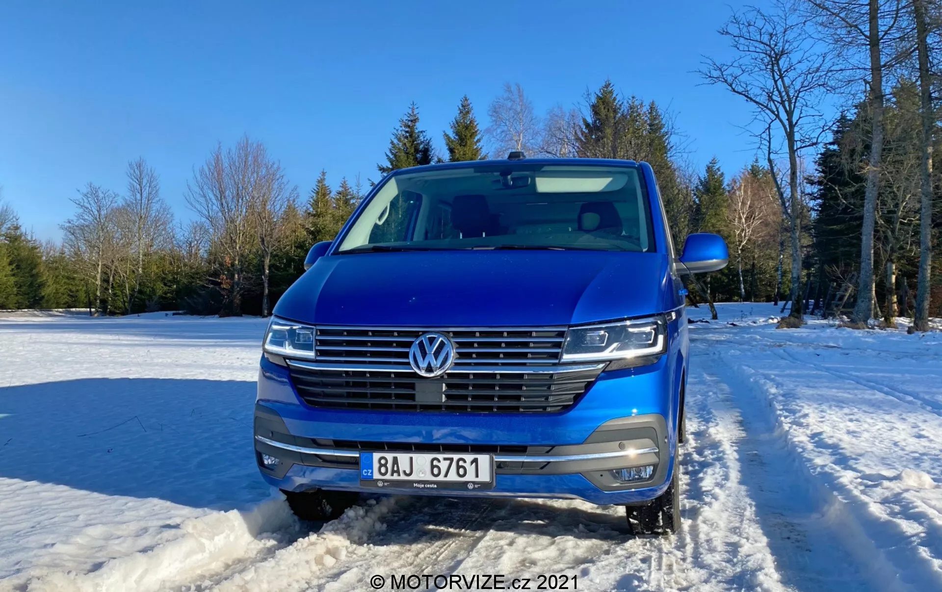 Frontalne ujęcie niebieskiego Volkswagena Transportera 2019 (T6-1 Multivan), ukazujące jego kratkę, reflektory i obszar maski. Pojazd znajduje się na zewnątrz na zaśnieżonym terenie z czystym niebem i drzewami w tle. Cechy samochodu obejmują emblem VW, poziomą kratkę z chromowanymi wykończeniami, reflektory LED, kamerę przednią, czujniki parkowania, srebrne relingi dachowe i częściowo widoczne boczne lusterko. Nowoczesny design z aerodynamicznym przodem, sfotografowany w ciągu dnia przy lekkim świetle słonecznym z prawej strony.