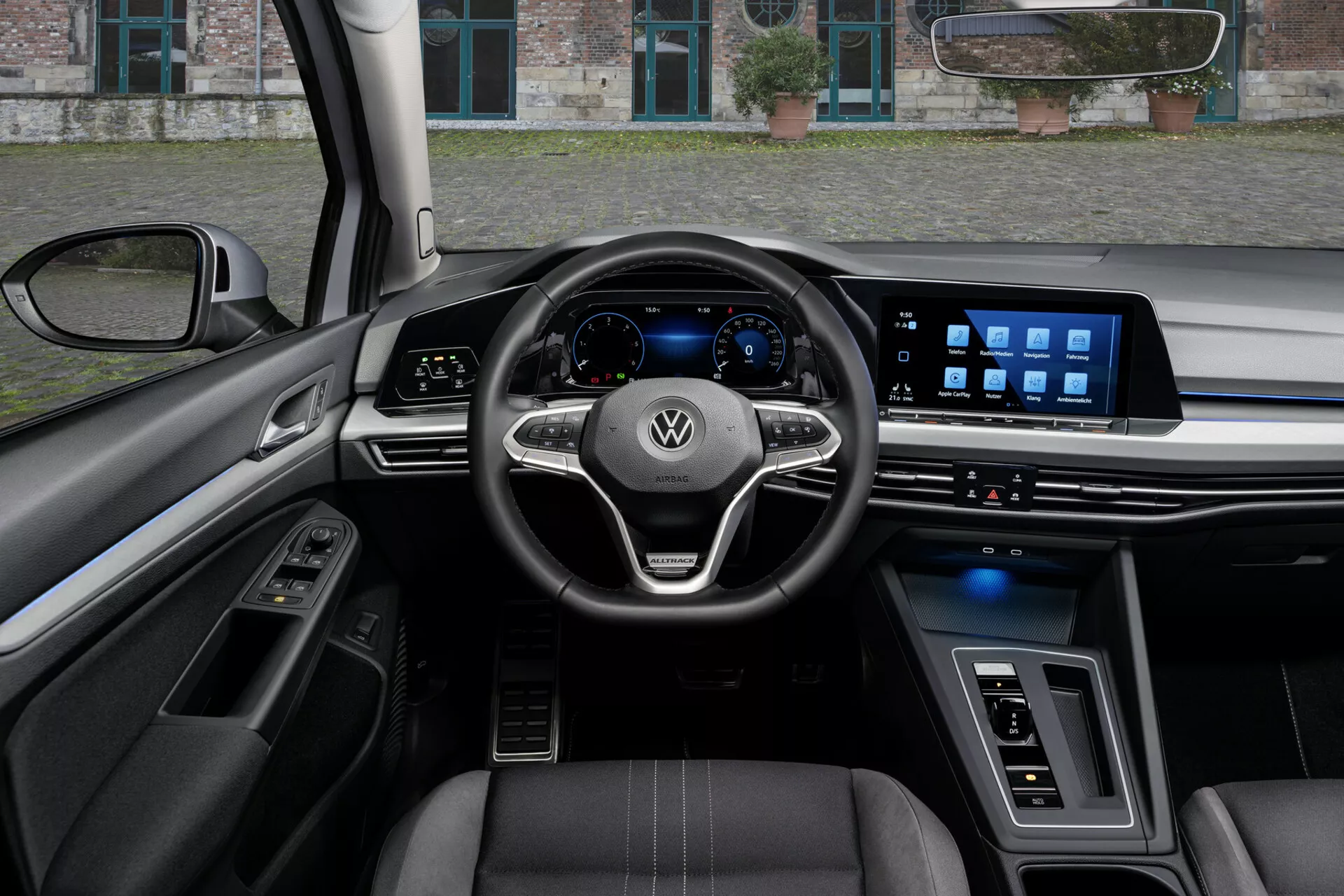 Widok wnętrza VW Golf Alltrack 2017 z perspektywy kierowcy, prezentujący kierownicę o trójramiennym wyglądzie z zamontowanymi sterowaniami i logo Volkswagen, cyfrowy zespół wskaźników i szeroki system infotainmentowy na ekranie dotykowym na desce rozdzielczej, uwydatniony zaawansowanymi funkcjami technologicznymi, fotele obite skórą z kontrastowym szyciem, i mieszanką luksusowych materiałów i oświetlenia ambientowego.