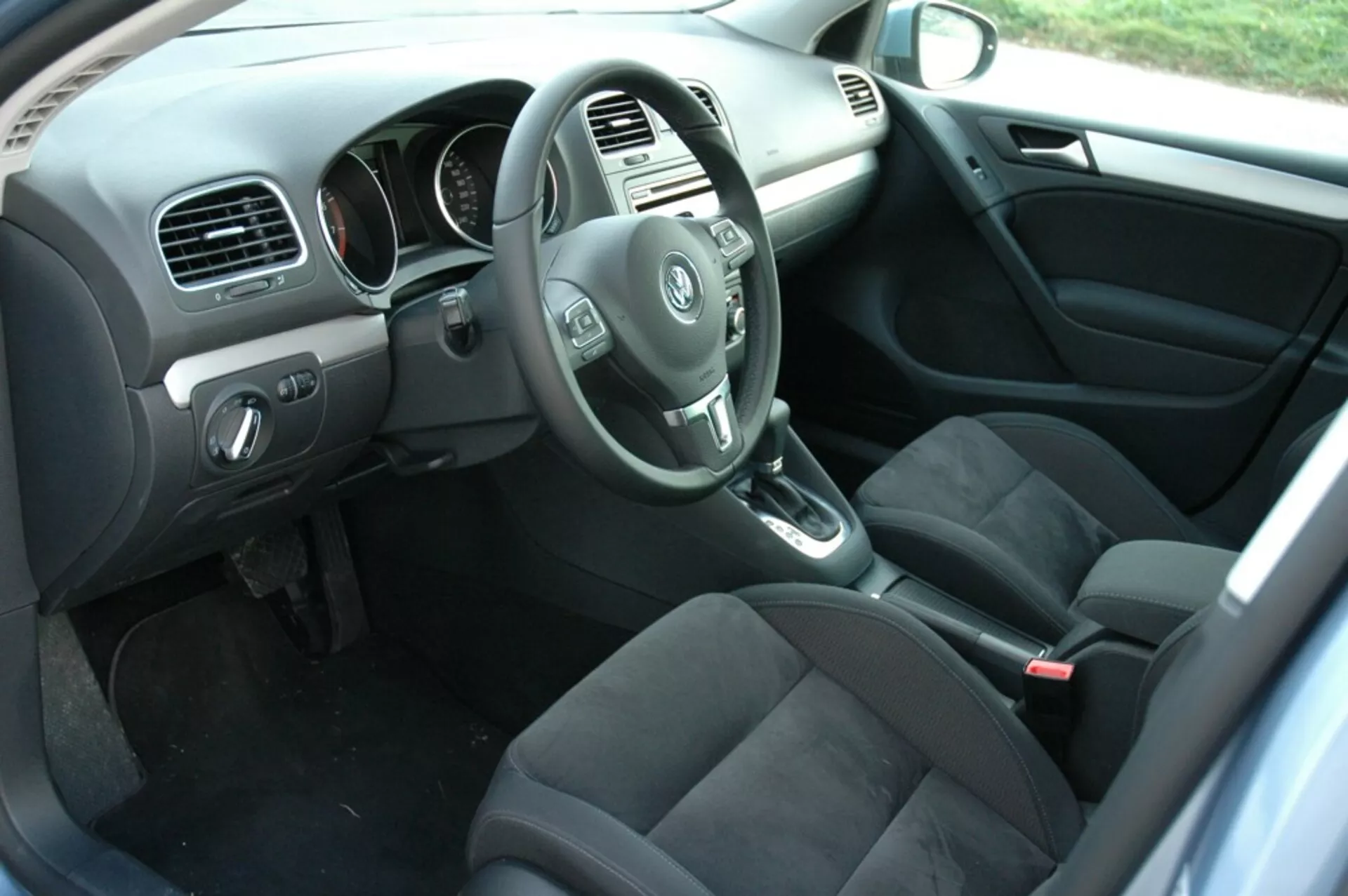 Wnętrze VW Golfa 2008 pokazujące stronę kierowcy z perspektywy tylnego pasażera z kierownicą, deską rozdzielczą, konsolą środkową, dźwignią zmiany biegów automatycznej skrzyni, ciemnymi tapicerowanymi siedzeniami, panelami sterowania na drzwiach kierowcy, wykładziną podłogową z matami i naturalnym oświetleniem