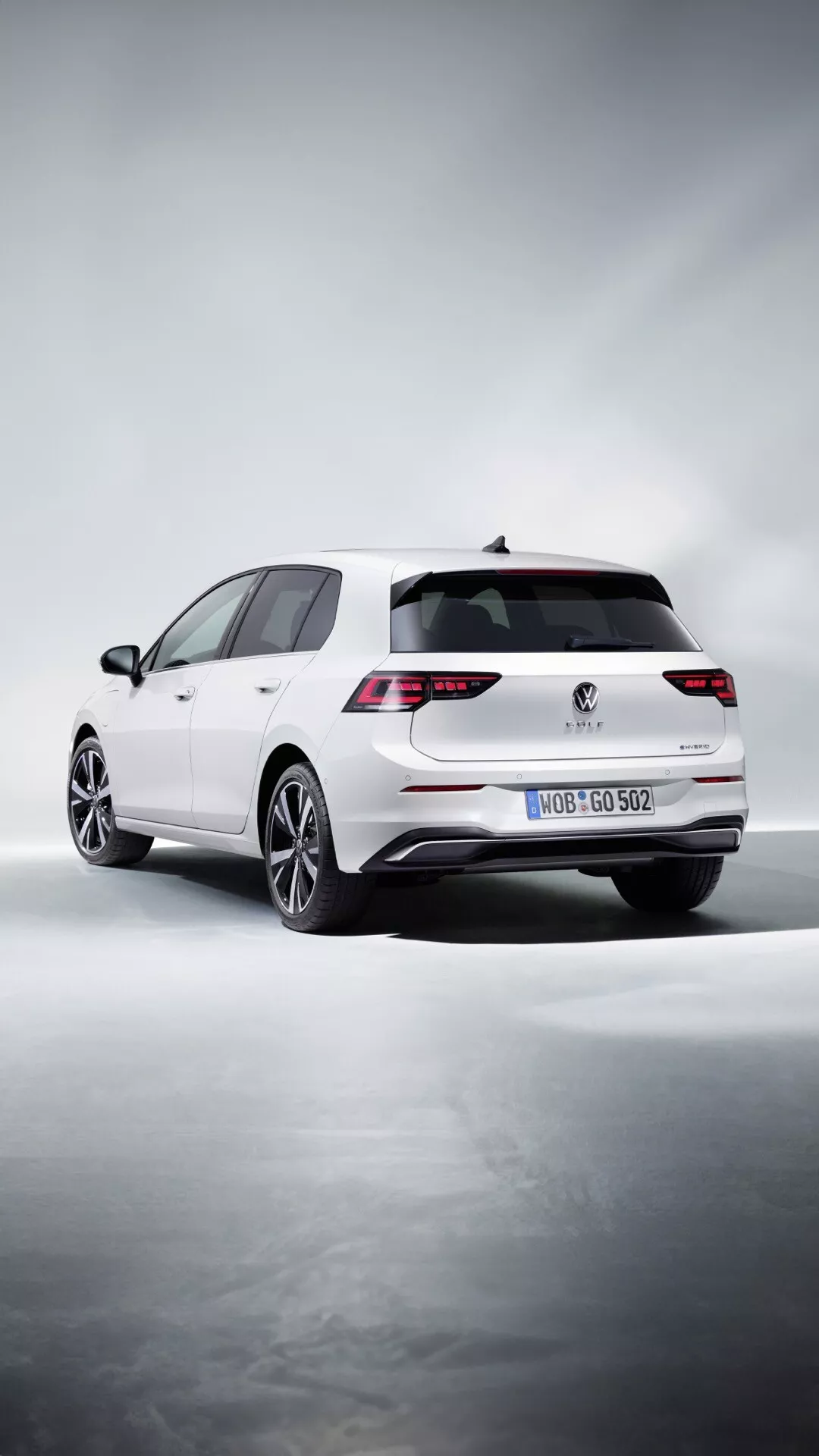 Biały hatchback Volkswagen Golf (VIII Facelift) 2024, perspektywa 3/4 tyłu prezentująca LEDowe tylne światła, podwójne rury wydechowe, alufelgi wieloszprychowe i spoiler dachowy na stylizowanym tle studia