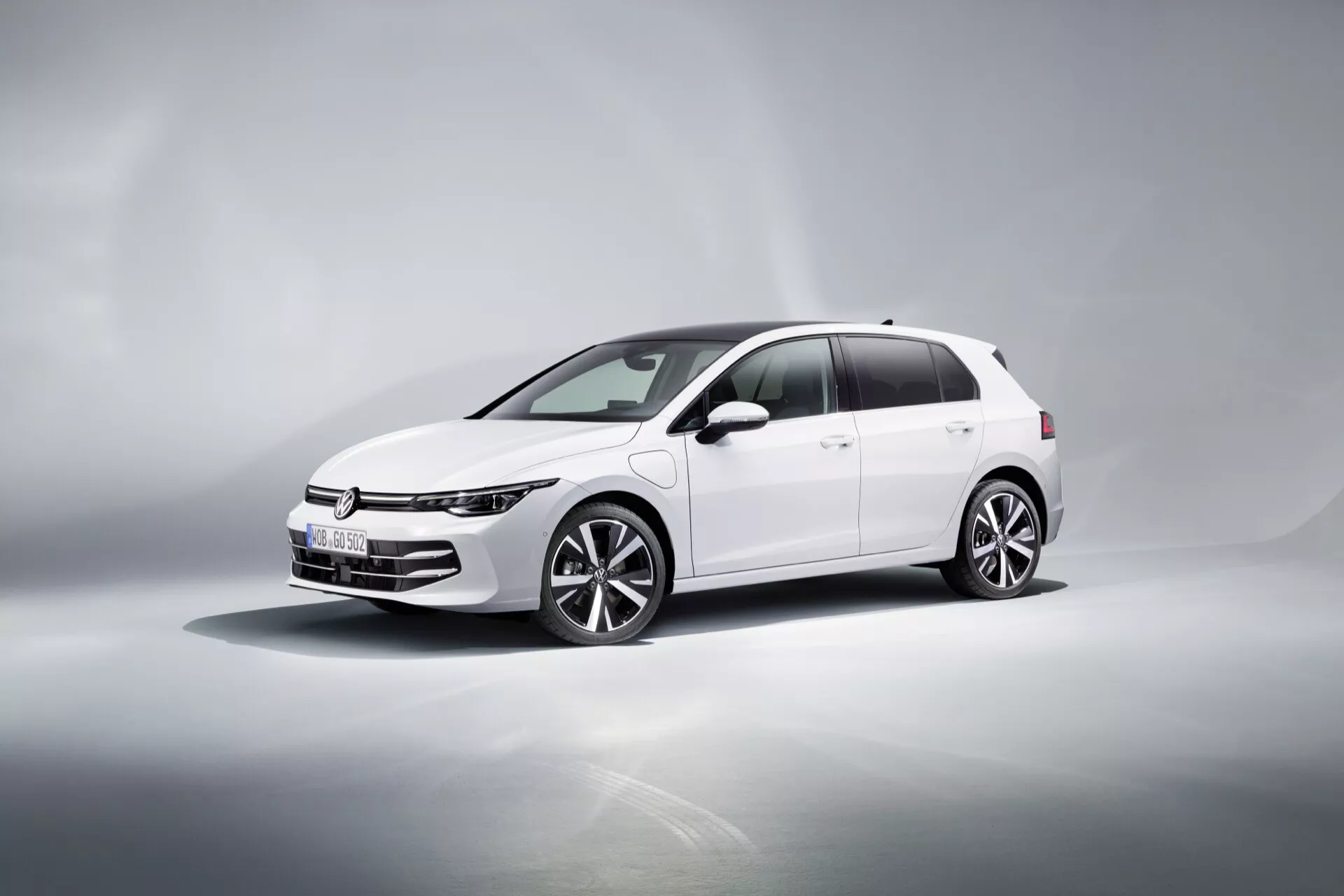 VW Golf (VIII-Facelift) 2024 w kolorze białym, prezentujący dynamiczny widok czołowo-boczny na odbijającej powierzchni z gradientowym tłem, podkreślający smukłe światła LED, poziomą atrapę chłodnicy, wieloramienne felgi aluminiowe, lusterka boczne z wbudowanymi kierunkowskazami oraz przyciemniane szyby.