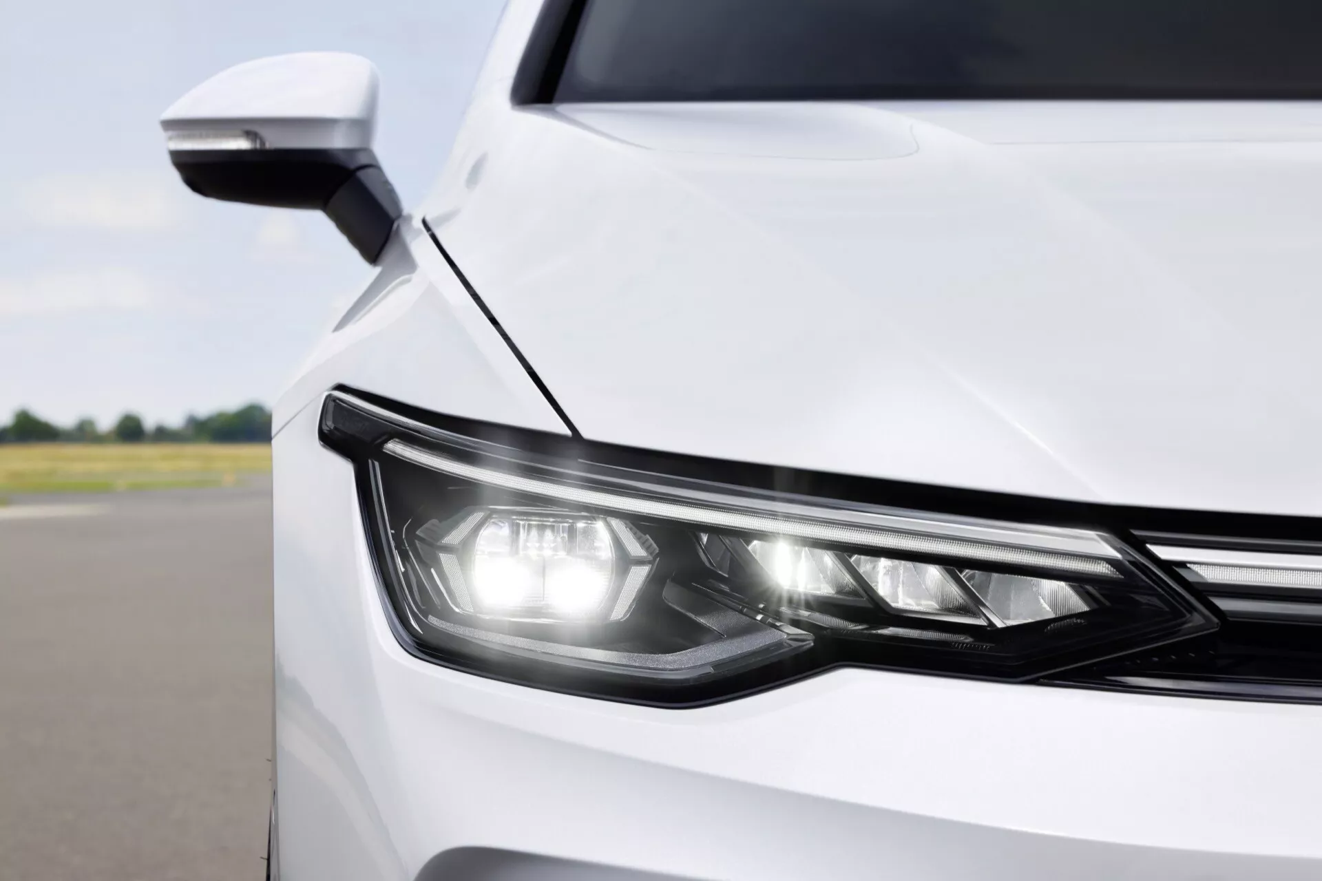 Zbliżenie na przedni narożnik białego VW Golfa VIII face liftingu 2024, prezentujące nowoczesny design i technologię reflektora LED i aerodynamicznego lusterka bocznego z wbudowanym kierunkowskazem, widok z przodu pod lekkim kątem