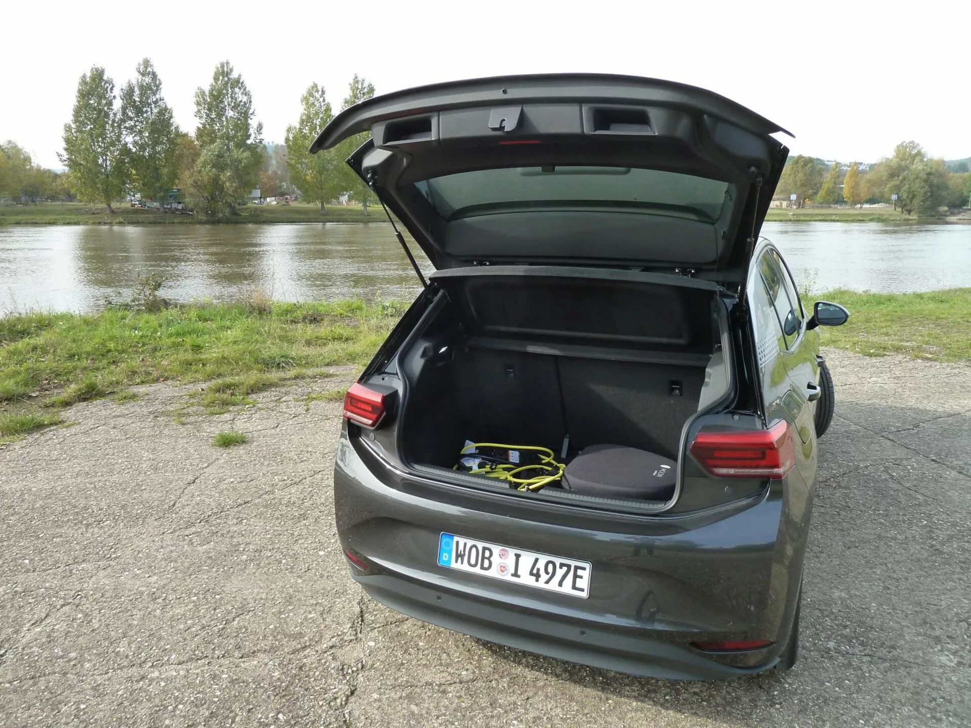 VW ID.3 z 2020 roku zaparkowany na żwirze obok wody z otwartym bagażnikiem pokazującym przestronne wnętrze i sprzęt sportowy, widok z tylu pod kątem trzech czwartych