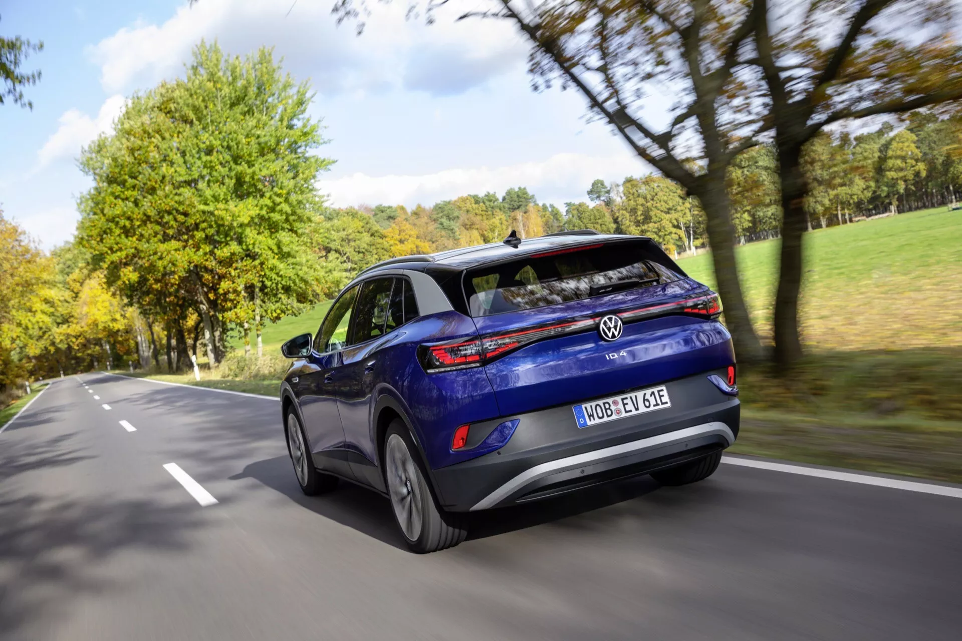 VW ID.4 1. generacji z 2020 roku w ruchu, uchwycony z tylnego kąta trzech czwartych prezentujący jego aerodynamiczny niebieski design hatchbacku, tylne światła LED i felgi aluminiowe na drodze dwupasmowej z efektem zamazania ruchu.
