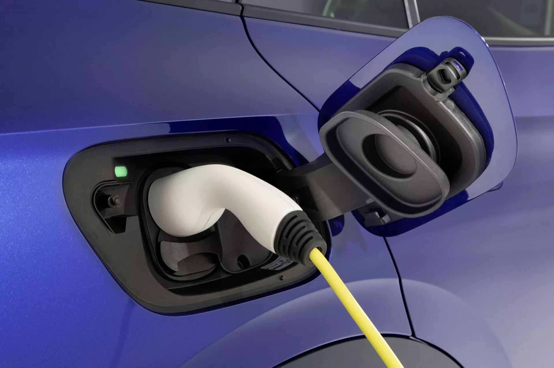 Zbliżenie na obszar portu ładowania elektrycznego pojazdu VW ID.4 2020 (1. generacja) z otwartą klapą wyświetlającą gniazdo i podłączony biały konektor, podkreślone zielonym światłem wskaźnika ładowania, na niebieskim nadwoziu samochodu.