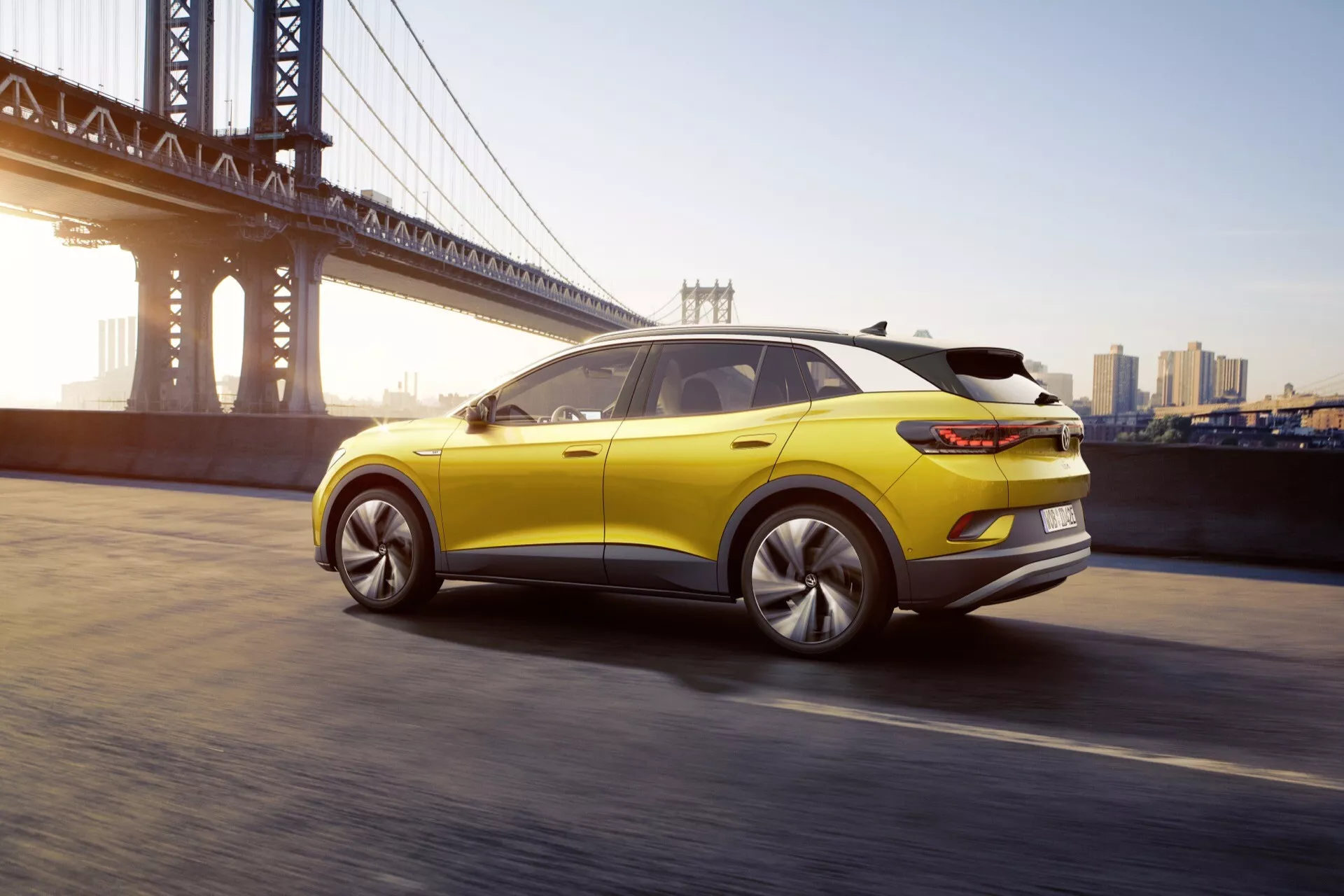 Dynamiczny widok z tyłu na trzy czwarte żółtego VW ID.4 z 2020 roku, podkreślający sportowy profil dachu, otaczające tylne światła LED i wieloramienne felgi aluminiowe, na tle miejskiego krajobrazu z wiszącym mostem i panoramą miasta o świcie lub zmierzchu.