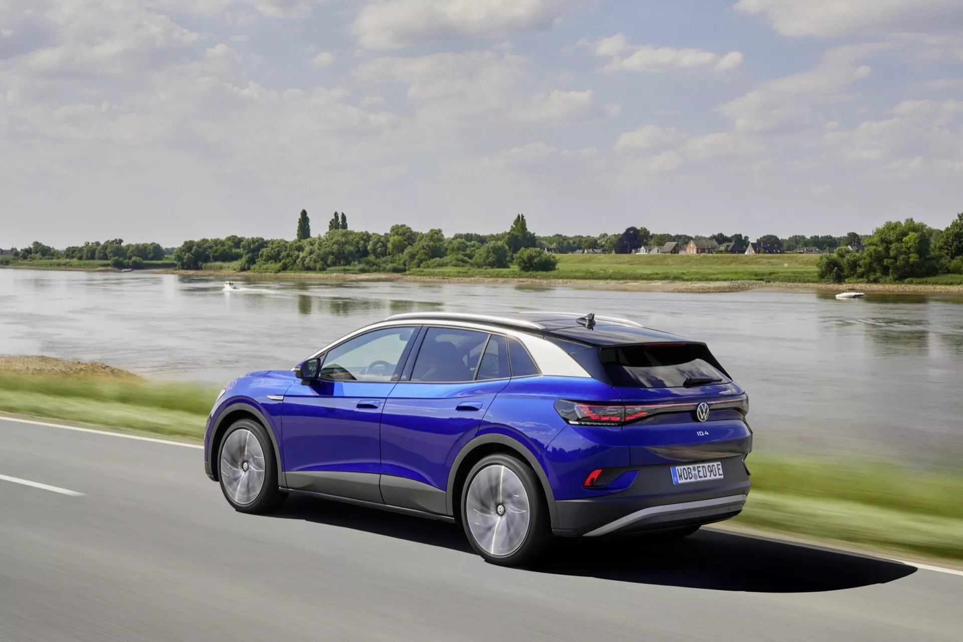 Dynamiczny boczny profil VW ID.4 1. generacji 2020 w żywym niebieskim kolorze, z opadającą liną dachu, sportowym spoilerm, felgami z lekkich stopów o wzorze wielkopręcikowym i zaawansowaną technologią LED, przekazujący ruch na drodze obok rzeki