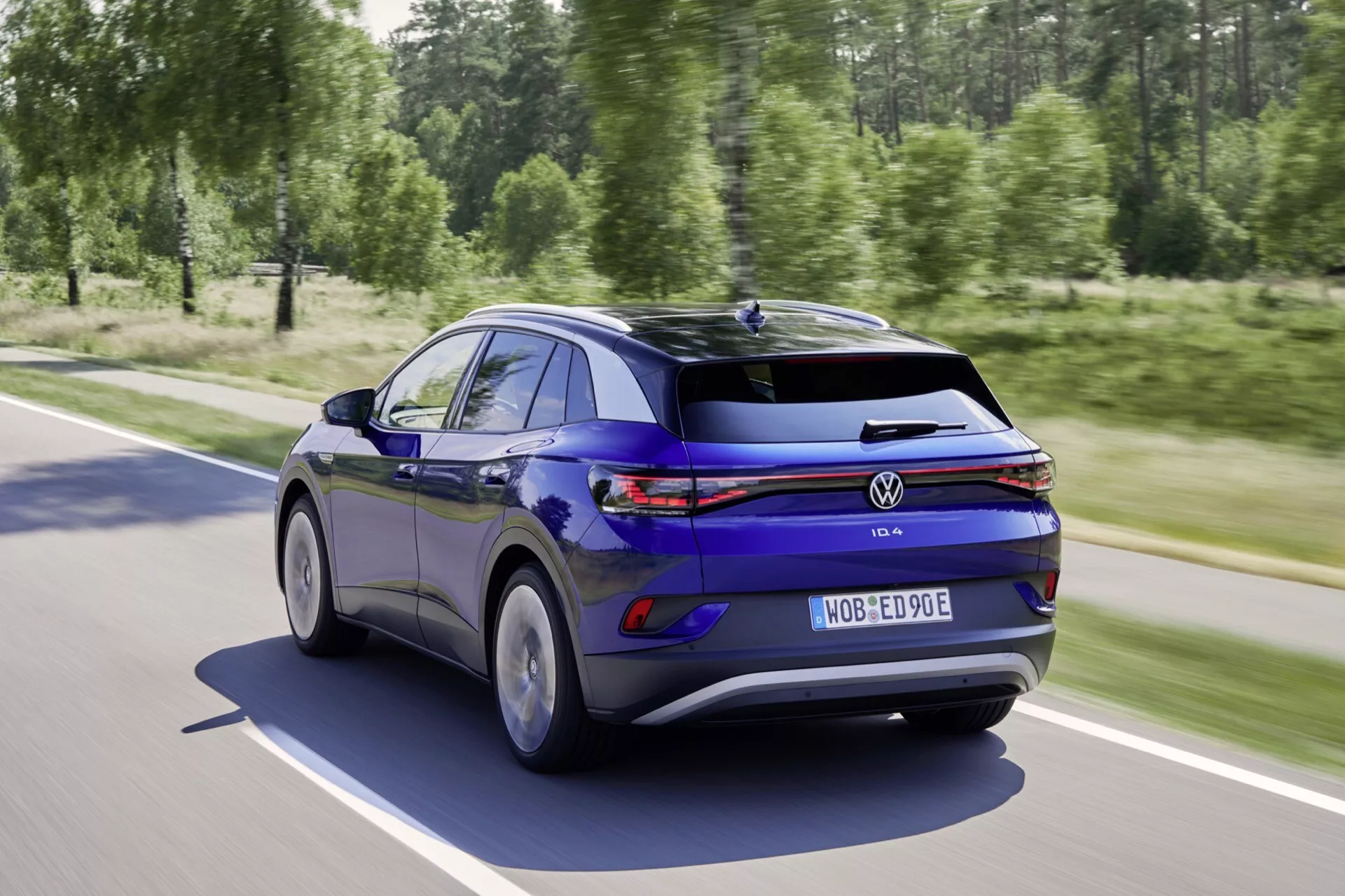 Hatchback Volkswagen ID.3 2020 w ruchu na słonecznej drodze, pokazany z tylnego ujęcia trzy czwarte podkreślającego jego elegancką niebieską farbę, ciągłe pasmo LED-owych świateł tylnych, aerodynamiczne koła i inteligentne funkcje technologiczne, w tym jazdę wspomaganą przez czujniki i efektywność elektryczną.