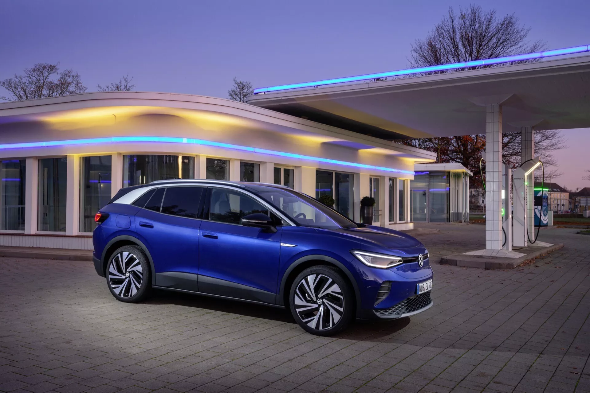 VW ID.4 1. generacji z 2020 roku zaparkowany na zewnątrz podczas zmierzchu, prezentujący metaliczny niebieski profil boczny na tle nowoczesnego budynku z ciepłym oświetleniem, podkreślający elegancki design SUV, oświetlenie LED i zaawansowaną aerodynamikę.