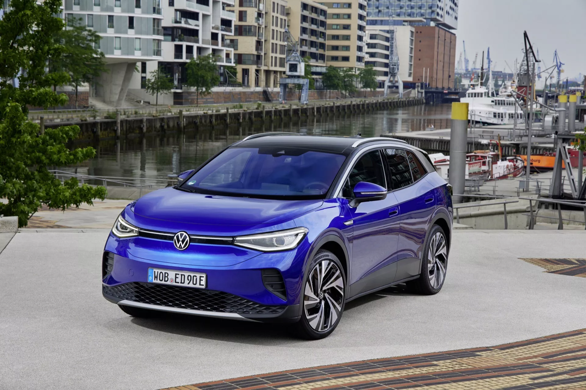 VW ID.4 1. generacji z 2020 roku z przedstawieniem grille, maski, lewego profilu boczego i części dachu, z naciskiem na stylizację reflektorów, projekt aerodynamiczny i technologię LED, na wzorzystym miejskim podjeździe na tle architektonicznym