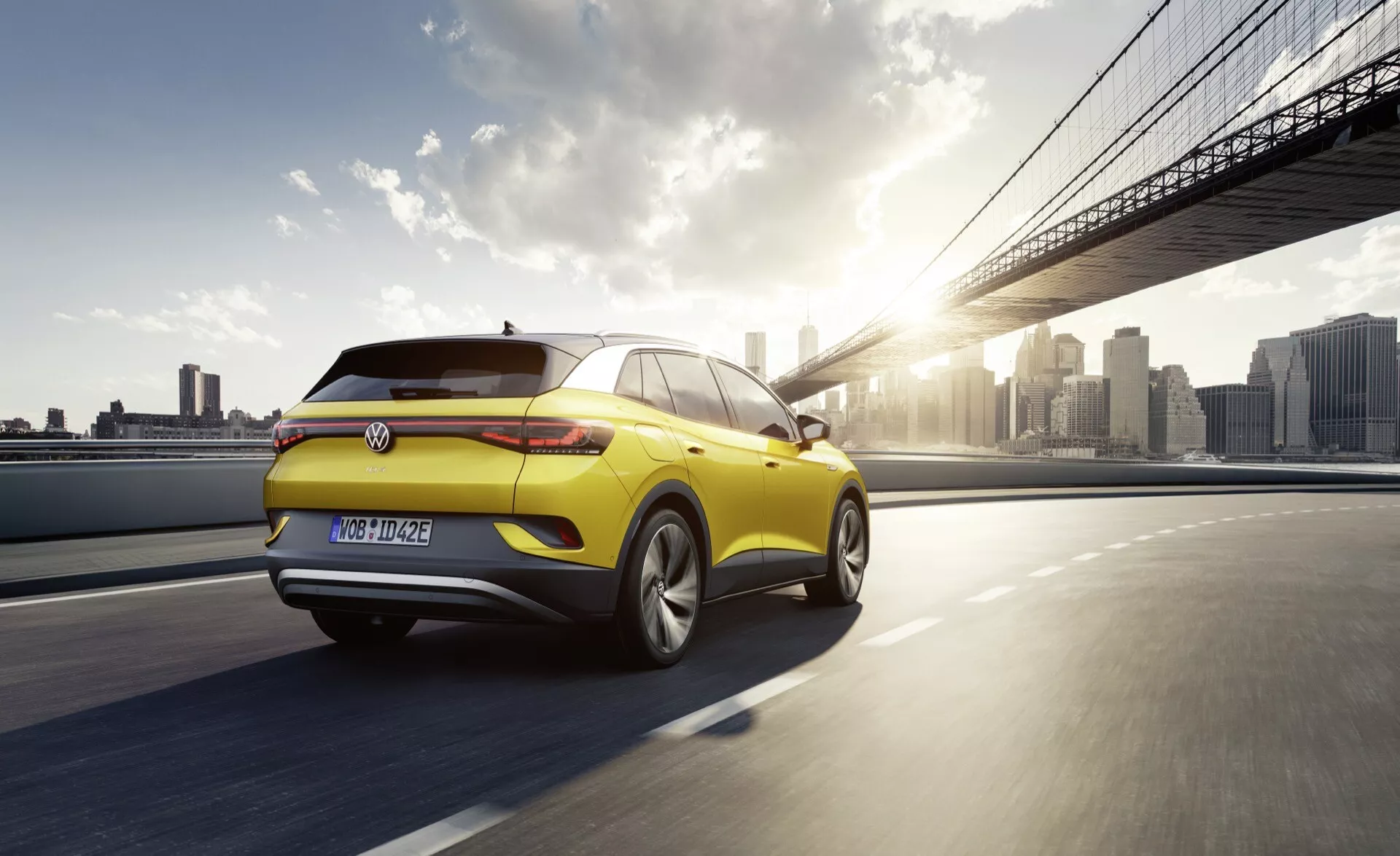 VW ID.4 1. generacji z 2020 roku w kolorze żółtym, jadący po moście o zachodzie słońca, prezentujący tylny i boczny profil z diodami LED, aerodynamicznym spojlerem oraz kołami z lekkich stopów