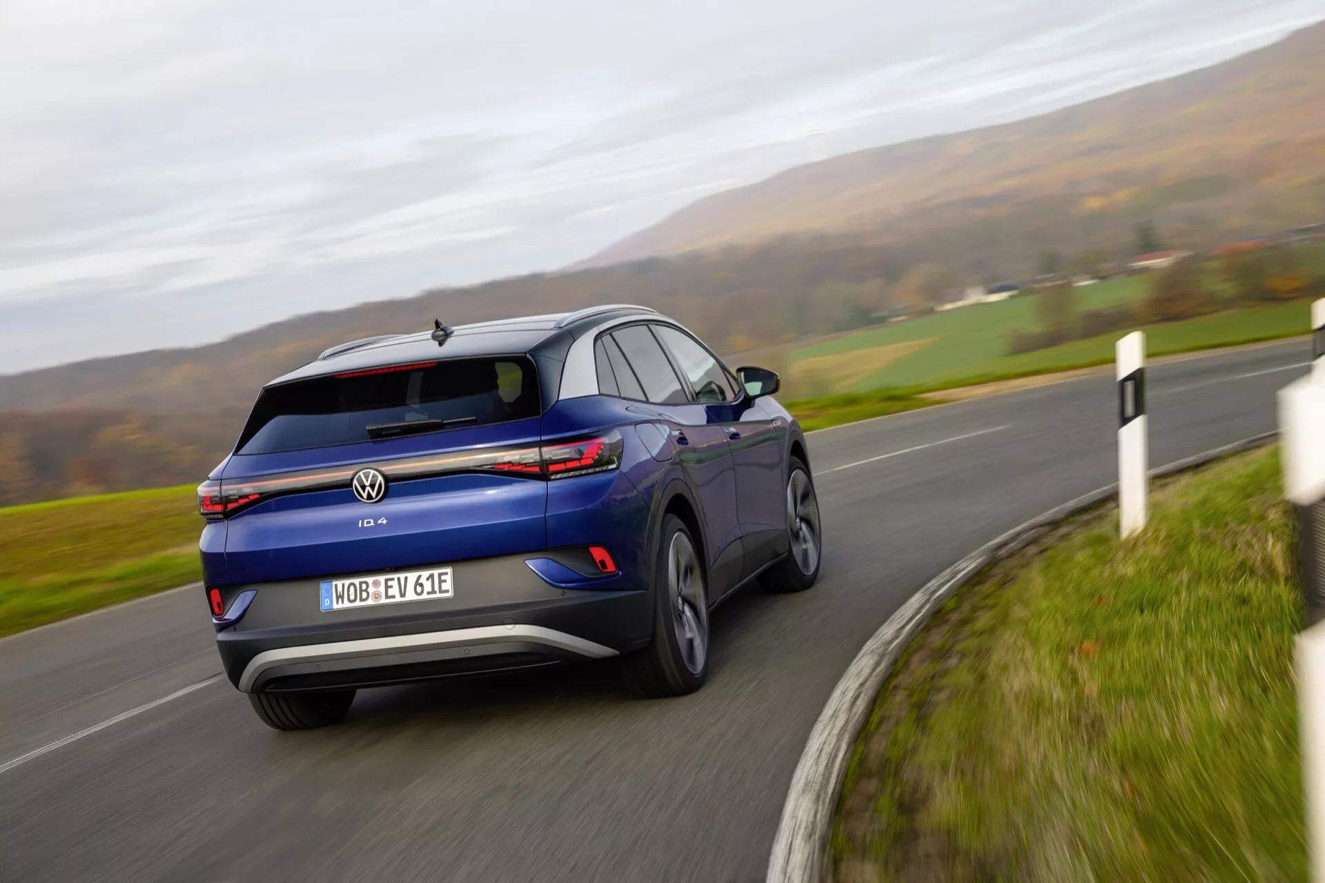 Dynamiczny widok z tyłu na trzy czwarte niebieskiego VW ID.4 2020 pokazujący rozmycie ruchu na krętej drodze, podkreślający elegancki design, LED-owe tylne światła i tablicę rejestracyjną kończącą się na 'E', co wskazuje na pojazd elektryczny