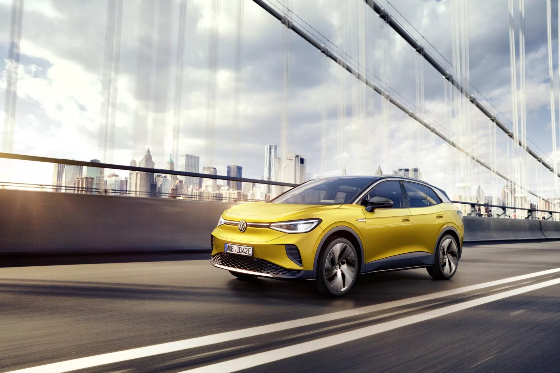 VW ID.4 1. generacja z 2020 w ruchu, pokazująca dynamiczny design i cechy technologiczne z niskiego kąta, trzy czwarte widoku z przodu po lewej stronie, z wyraźnym żółtym kolorem pojazdu, nowoczesnymi felgami aluminiowymi i potencjalnym ADAS na rozmytym miejskim tle