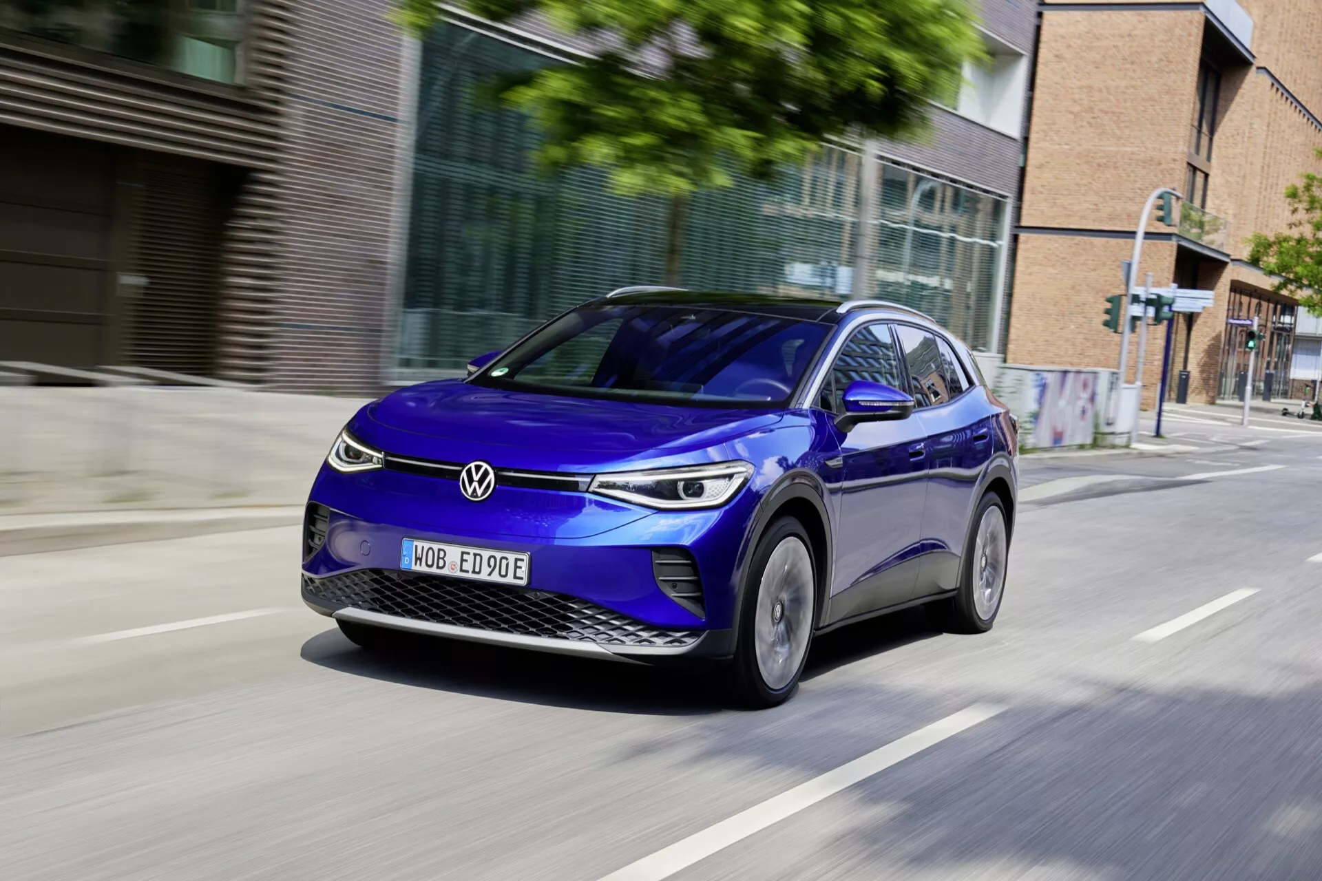 Dynamiczne zdjęcie niebieskiego elektrycznego hatchbacka czterodrzwiowego VW ID.4 1 generacji z 2020 roku w ruchu na miejskiej drodze, przedstawiające widok z przodu z perspektywy trzech czwartych, podkreślające nowoczesny design, reflektory LED, światła do jazdy dziennej i aerodynamiczne felgi aluminiowe