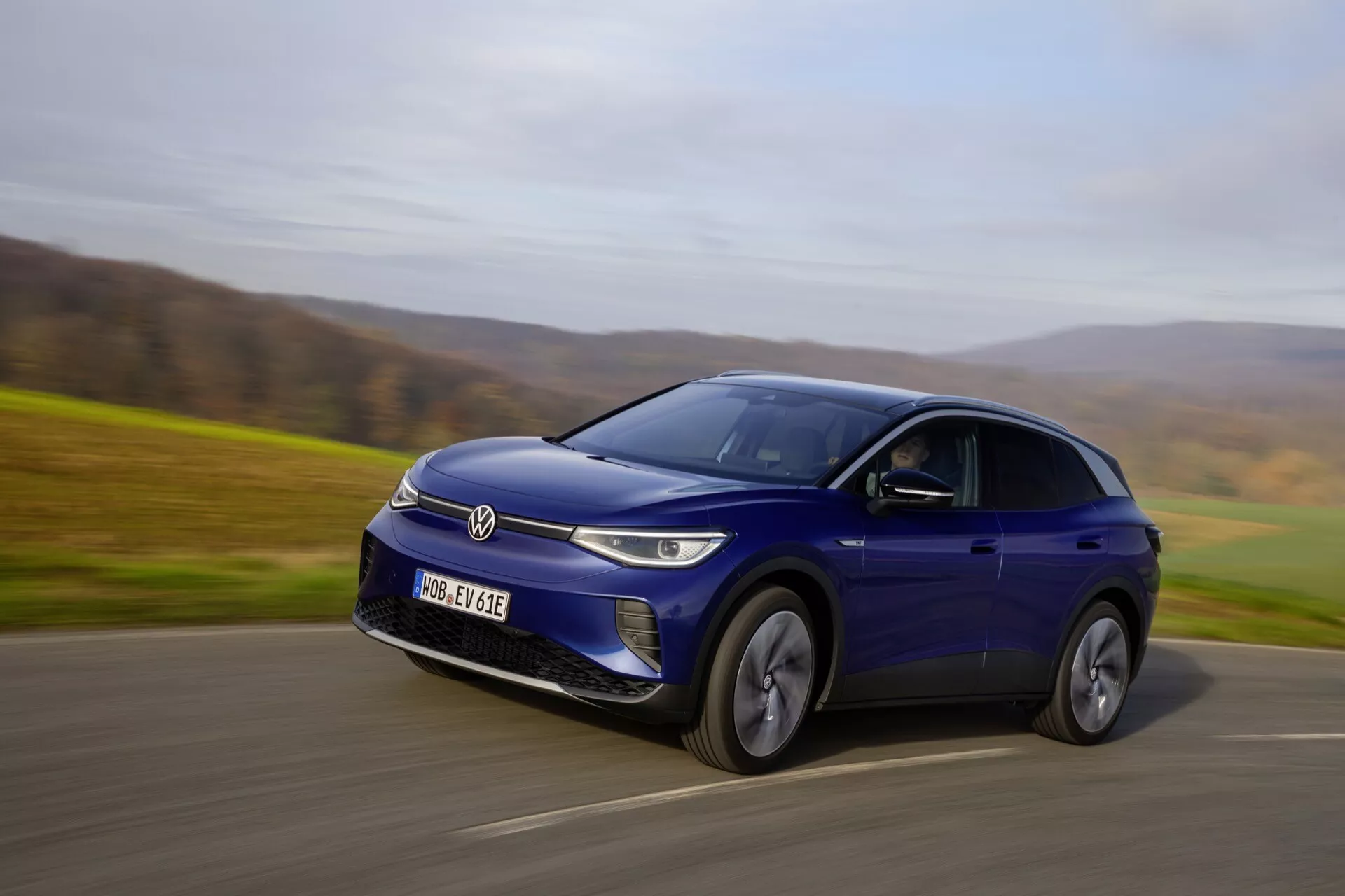 Niebieski elektryczny crossover SUV Volkswagen ID.4 w ruchu na wiejskiej drodze, uchwycony z perspektywy przedniej trzy czwarte, podkreślające jego dynamiczny ruch i cechy konstrukcyjne takie jak zamknięta atrapa z reflektorami LED, felgi aluminiowe i smukła, sportowa sylwetka pod metalicznym niebieskim wykończeniem.