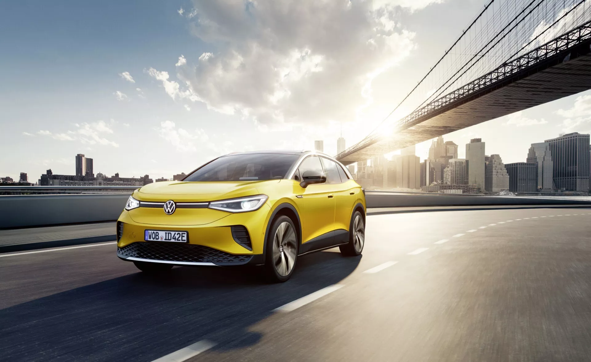 Dynamiczne zdjęcie jaskrawożółtego samochodu elektrycznego Volkswagen ID4 2020 w ruchu na drodze, prezentującego jego sportowy profil i nowoczesne cechy projektowania, takie jak światła LED, aerodynamiczna karoseria i koła ze stopu, na tle miejskiej linii horyzontu o wschodzie lub zachodzie słońca.