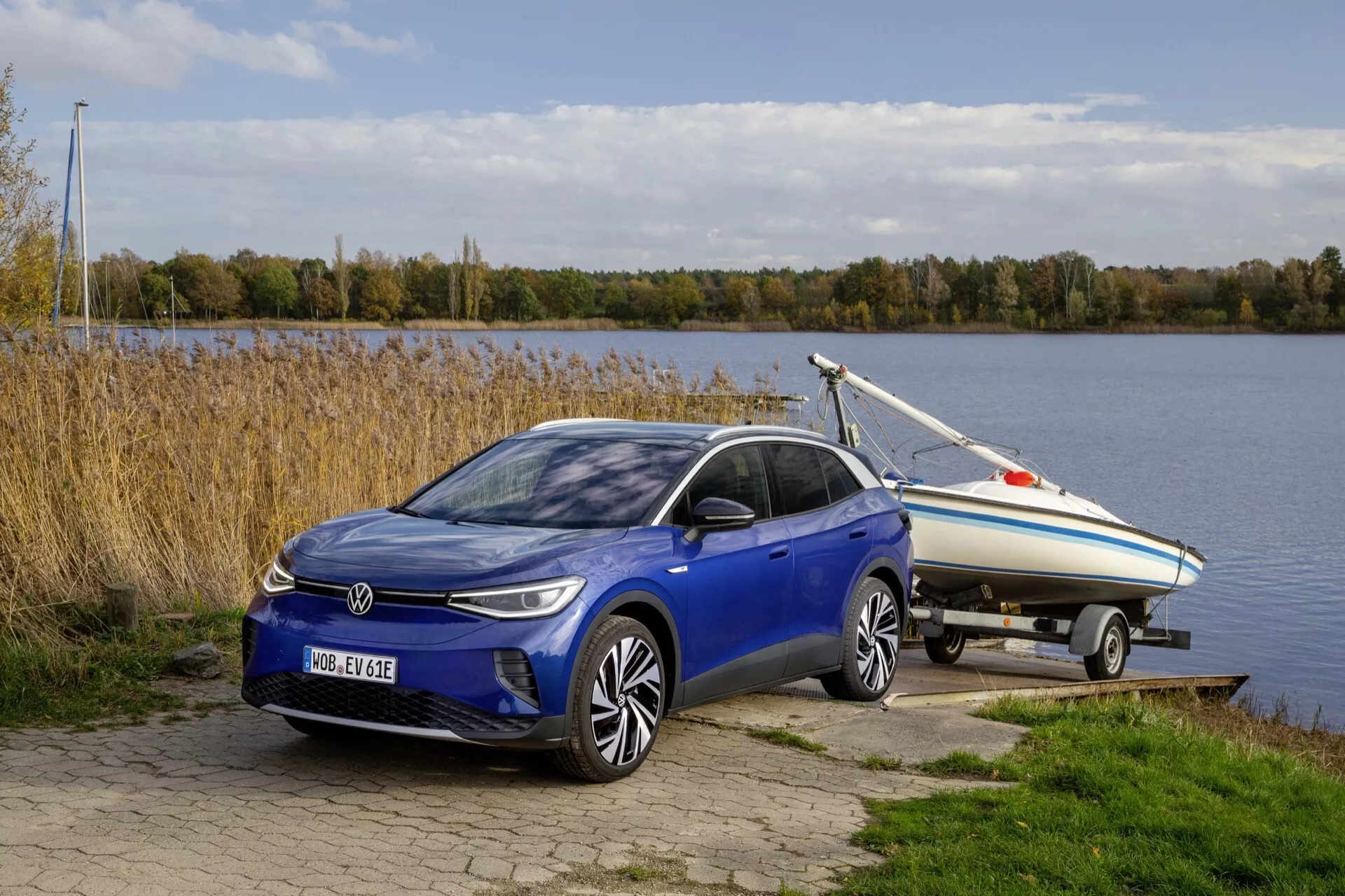 VW ID.4 1. generacji z 2020 roku na zewnątrz, holujący łódź, podkreślony z perspektywy ¾ frontalnej ukazującej jego elegancką, elektryczną lub hybrydową konstrukcję hatchback, reflektory LED, felgi aluminiowe, i umieszczony w pobliżu akwenu z spokojnym naturalnym tłem