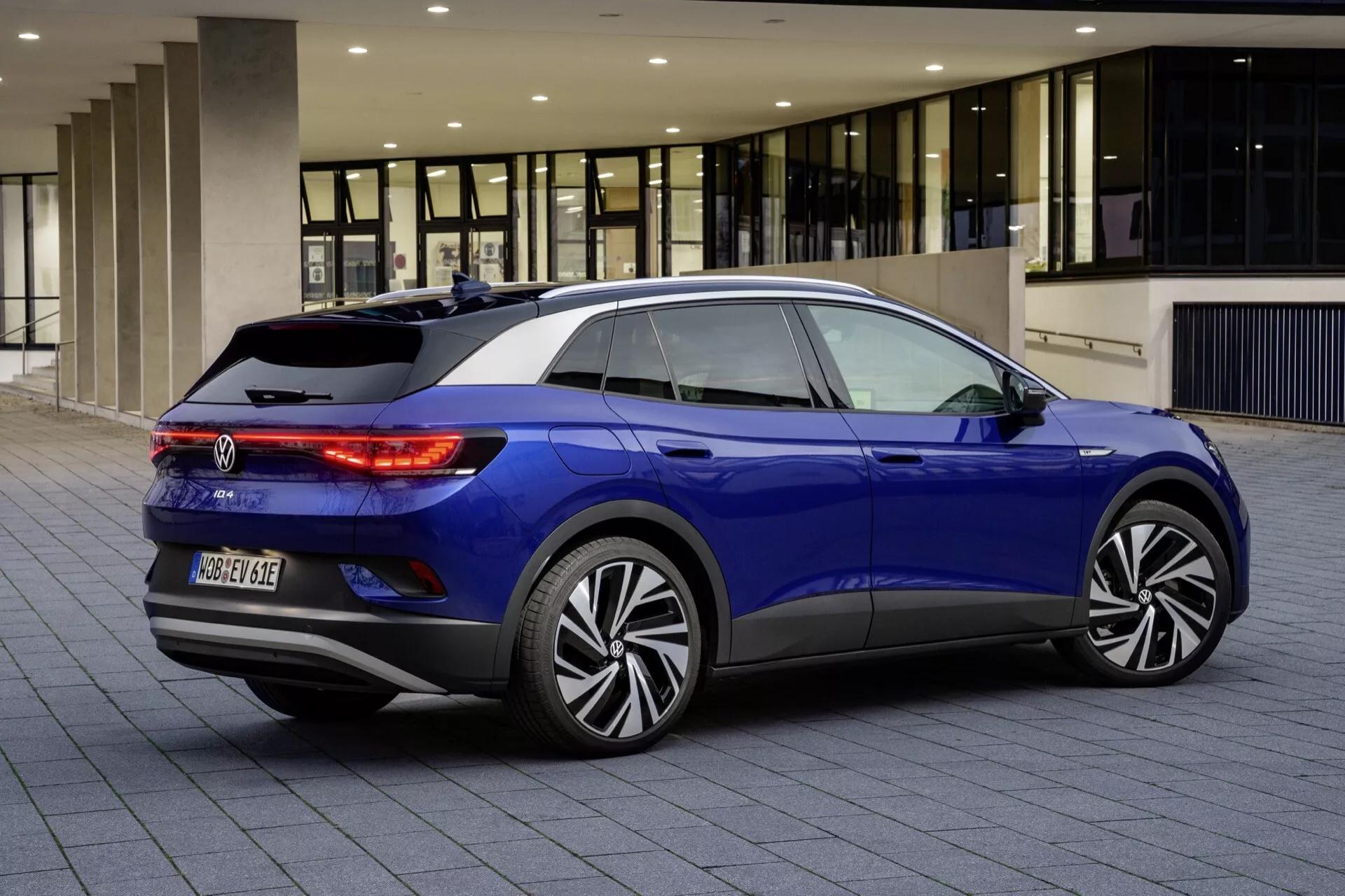 Niebieski hatchback VW ID.4 1. generacji 2020, widok z tyłu i prawego boku na utwardzonej nawierzchni, ukazujący diody LED w tylnych światłach, emblemat modelu, wieloramienne felgi aluminiowe, tylny zderzak z czujnikami parkowania, opadającą linię dachu ze spoilerem, tylną szybę z ogrzewaniem i wycieraczką oraz przyciemniane szyby boczne.