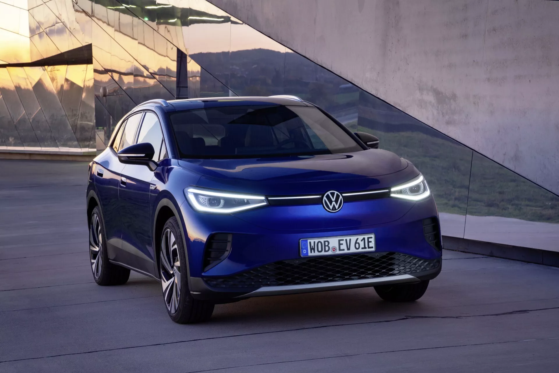 Niebieski VW ID.4 pierwszej generacji z 2020 roku, pokazany z niskiego kąta trzy czwarte z przodu, prezentujący lewą stronę pojazdu, nowoczesna grill zamknięty, emblemat producenta, smukłe reflektory LED z światłami do jazdy dziennej, aerodynamiczne felgi aluminiowe, zintegrowane kierunkowskazy w lusterkach bocznych, klapka portu ładowania po stronie kierowcy oraz rzeźbiona karoseria dla lepszej aerodynamiki i stylu.
