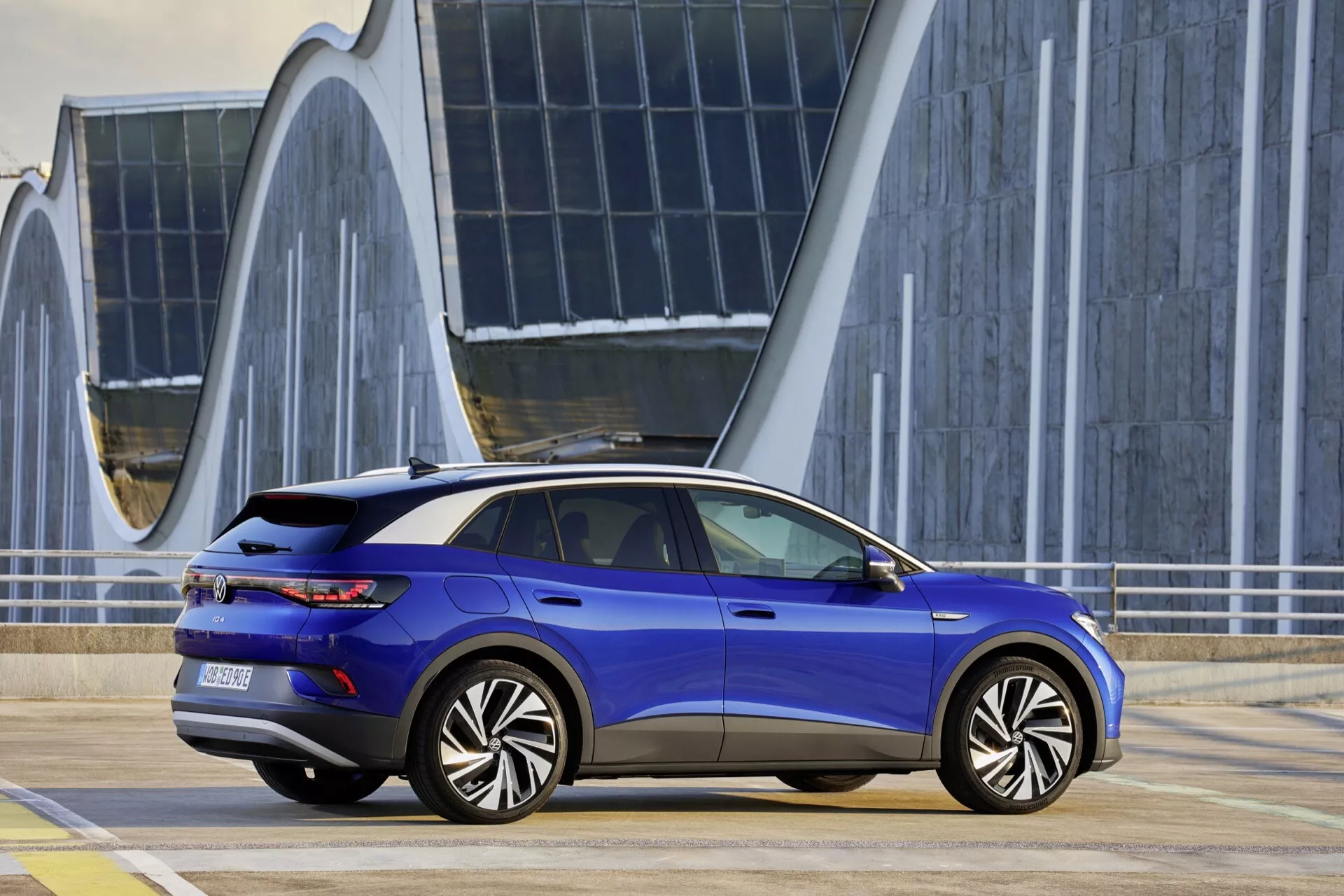VW ID.4 1. generacji 2020 w metalicznie niebieskim kolorze, prezentujący stylowy profil boczny i tylny z aerodynamicznym wzornictwem, klamki drzwiowe w linii z nadwoziem i zintegrowany pas świetlny, zaparkowany na wybrukowanym terenie z nowoczesną architekturą w tle