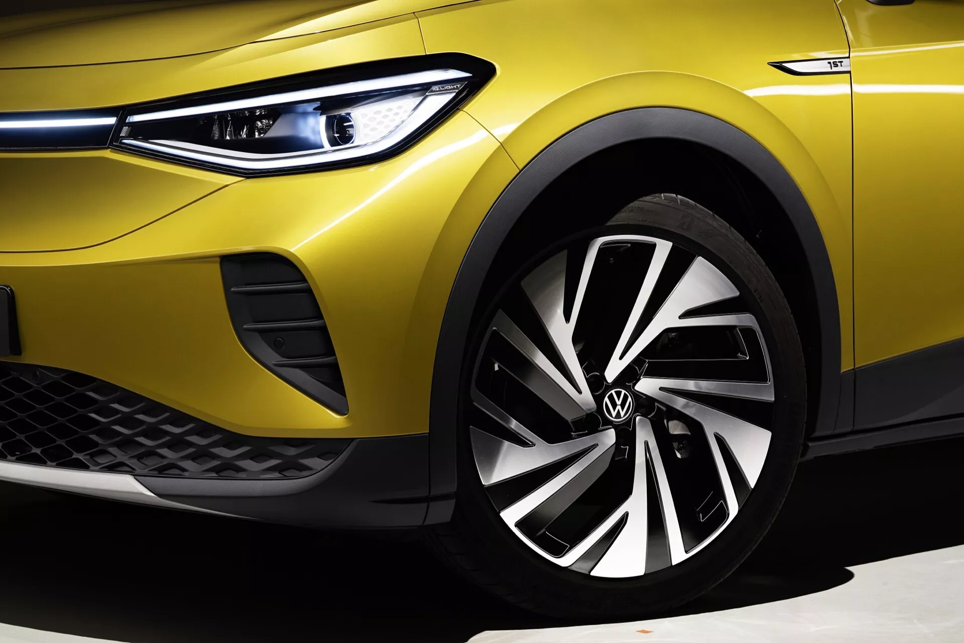 Zbliżenie na VW ID.4 1. generacji z 2020 roku przedstawiające jaskrawożółty lakier, reflektory LED z funkcją świateł do jazdy dziennej, sportowy przedni zderzak z wzorem plaster miodu, felgi aluminiowe o wielu szprychach i niskoprofilowe opony pod jasnym oświetleniem