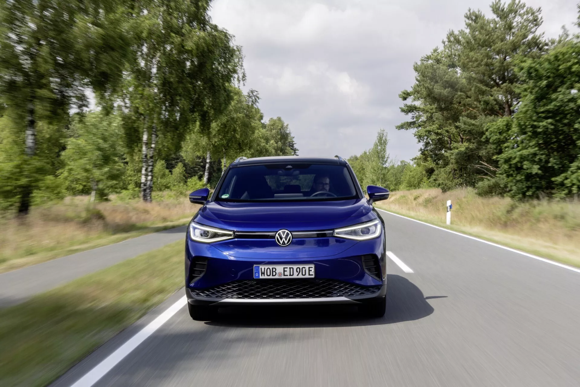 Czołowa perspektywa niebieskiego VW ID4 2020 jadącego drogą otoczoną zielenią, podkreślająca jego nowoczesny design elektryczny, aerodynamiczne linie oraz zaawansowane reflektory LED.