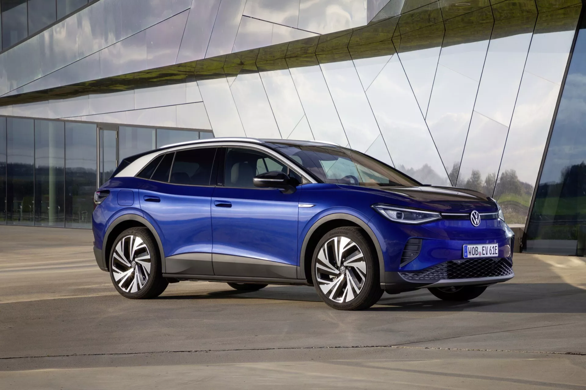 VW ID.4 1. generacji, rok 2020, w metalicznie niebieskim kolorze, zaparkowany na zewnątrz, o eleganckim wyglądzie z pochyłym dachem, reflektorami LED i wieloramionowymi kołami ze stopów, na tle nowoczesnego budynku