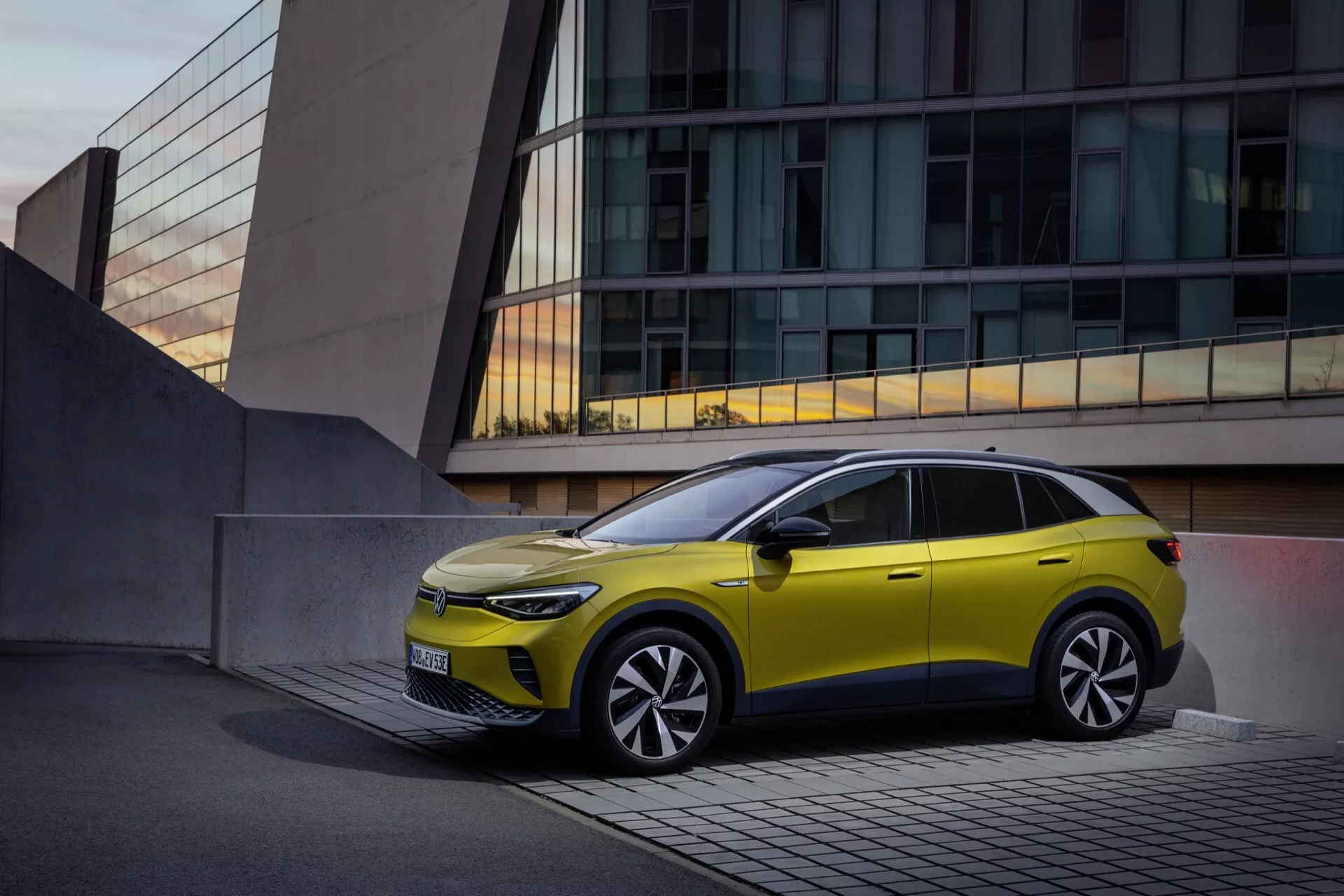 VW ID.4 1. generacji z 2020 roku w jasnożółtym kolorze zaparkowany przed współczesnym budynkiem, pokazujący smukłe reflektory LED, minimalną kratkę świadczącą o elektrycznym lub hybrydowym projekcie, rzeźbioną, aerodynamiczną maskę, duże koła ze stopu z wieloramienneym wzorem, zintegrowane kierunkowskazy w lusterkach bocznych, wyrazistą linię charakteru, czarną obwódkę okna z dachem przesuwnym, i unikatowy design okna drzwi tylnych odzwierciedlający nowoczesną technologię motoryzacyjną i cechy premium na tle spokojnego miejskiego krajobrazu.