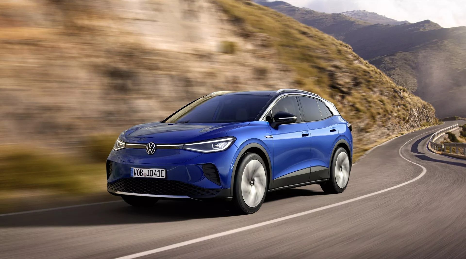 Dynamiczny widok niebieskiego VW ID4 1. generacji z 2020 roku, prezentujący profile przednie i boczne z reflektorami LED, aerodynamicznym designem i kołami ze stopów na zakrzywionej drodze, sugerujący zaawansowaną technologię i możliwości elektryczne lub hybrydowe
